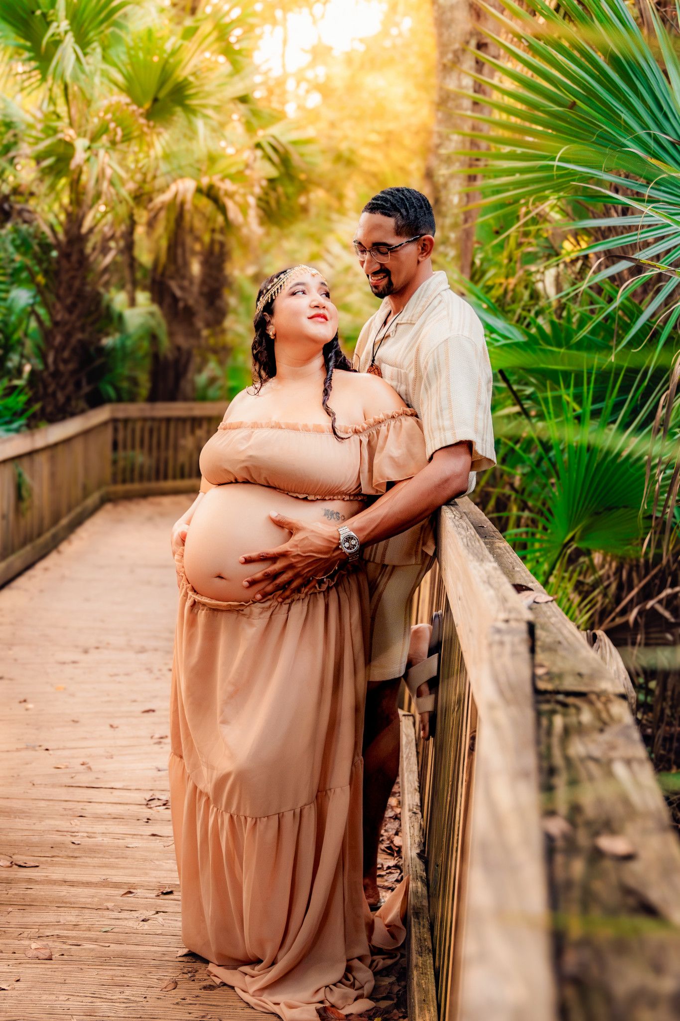 Pregnant woman in peach dress with partner on wooden boardwalk surrounded by tropical palm trees during maternity photoshoot.