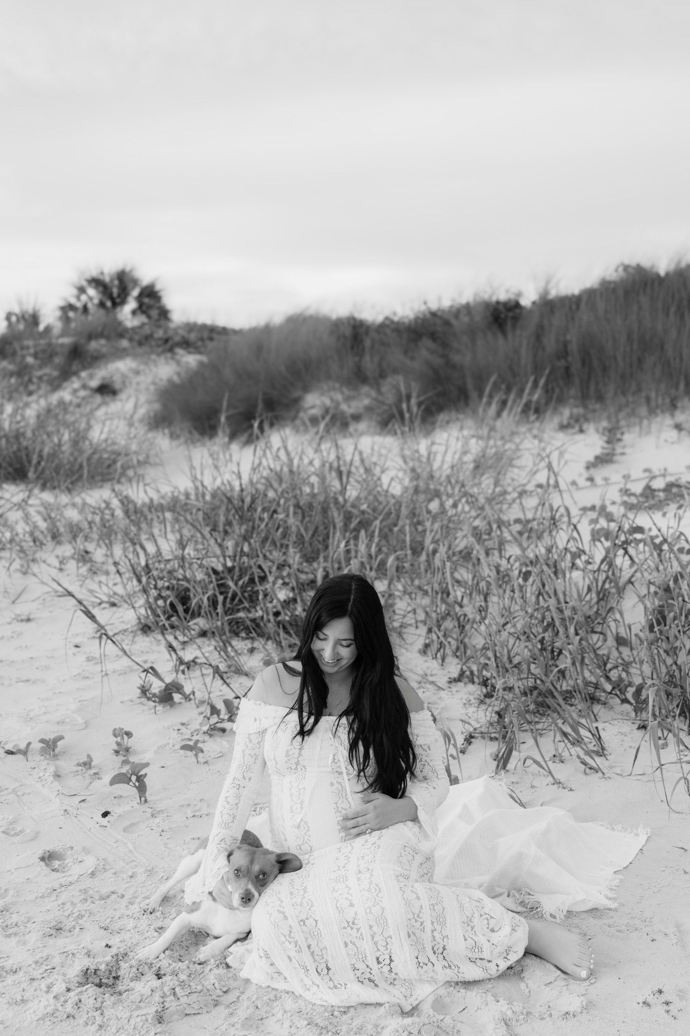 Black and white photo of a woman in a white lace dress sitting on sand dunes with a small dog.