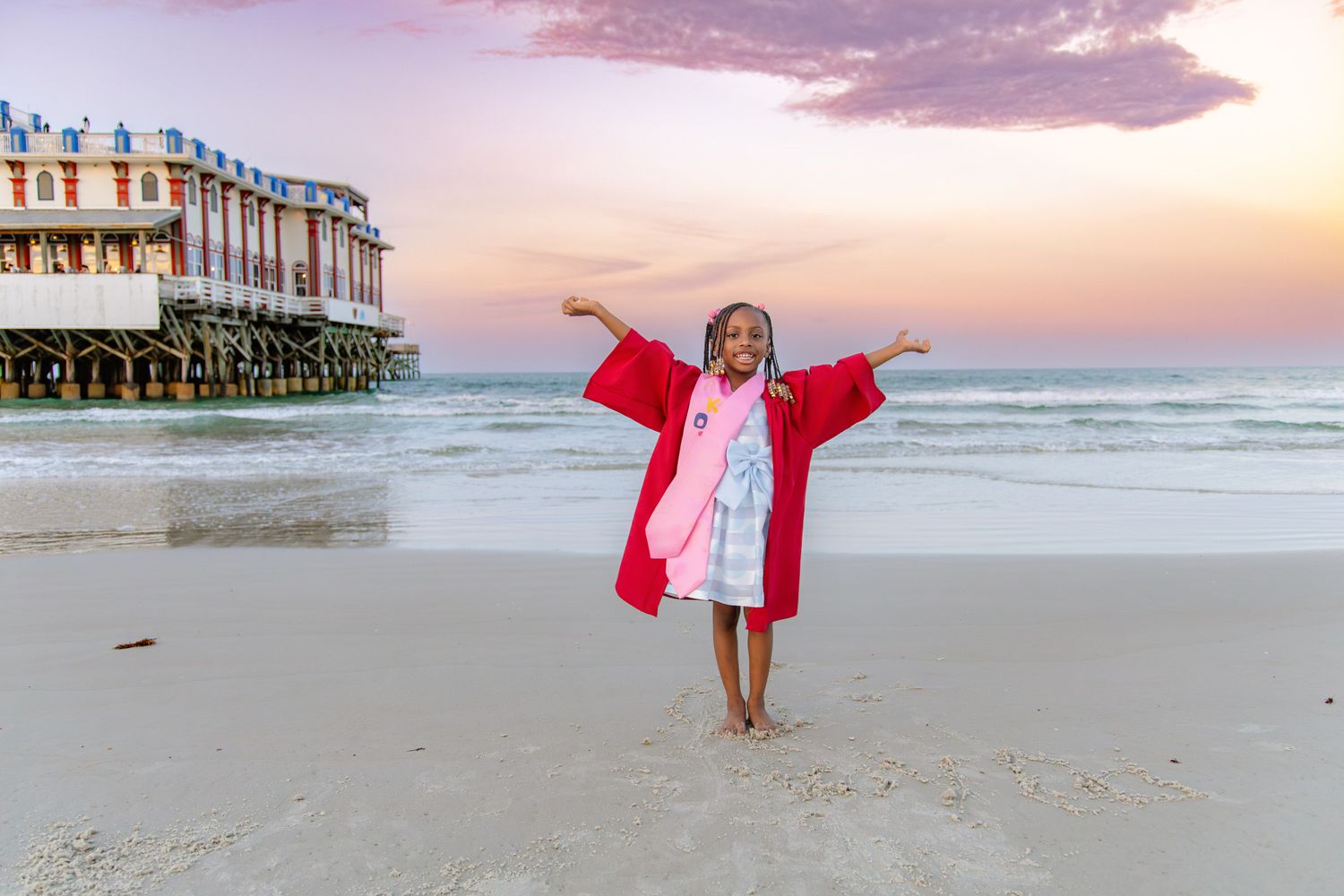 A joyful child in a red hooded towel poses on a sandy beach at sunset with a pier in the background.