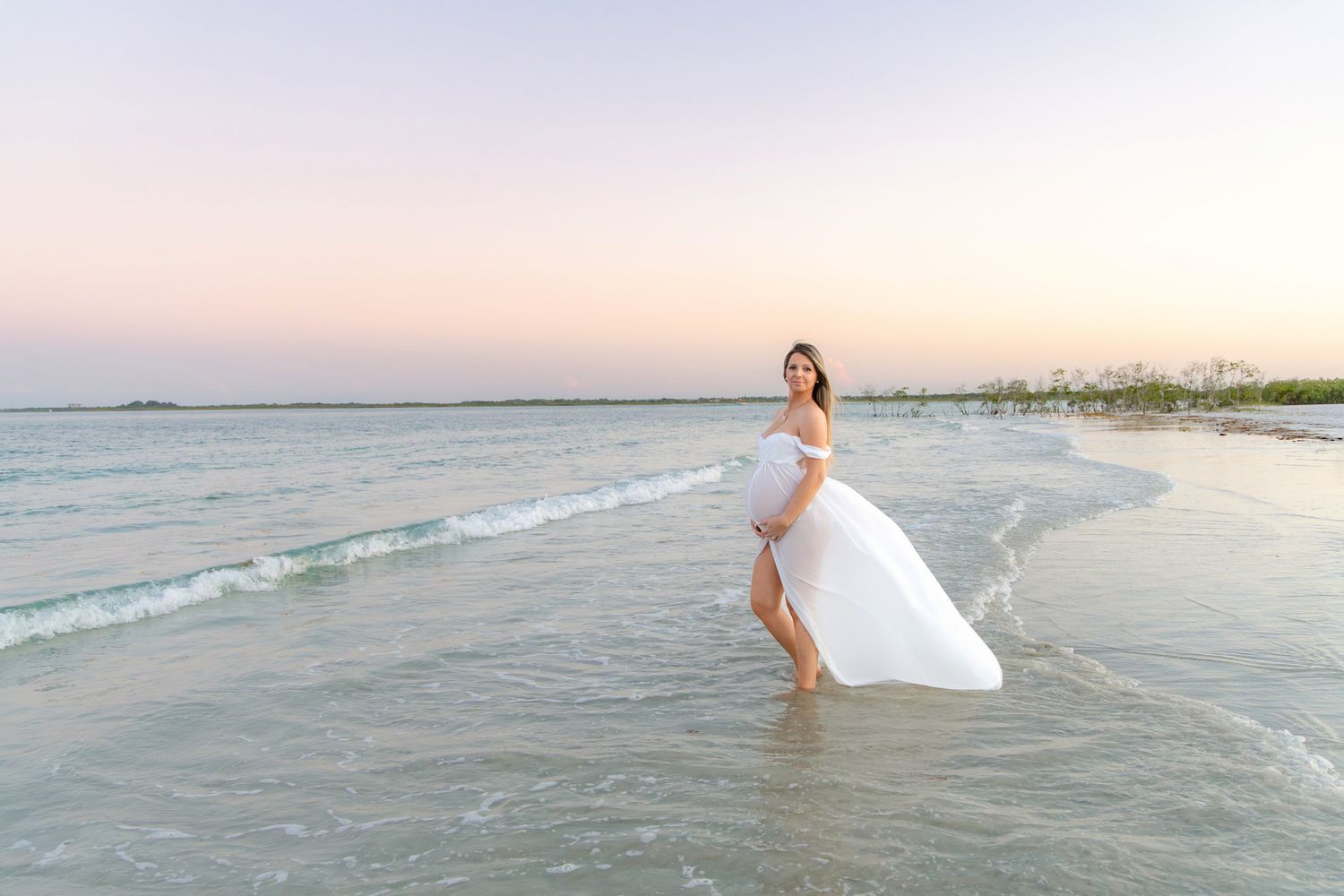 Pregnant woman in white flowing dress standing in shallow ocean waves at a pastel pink and blue sunset beach.
