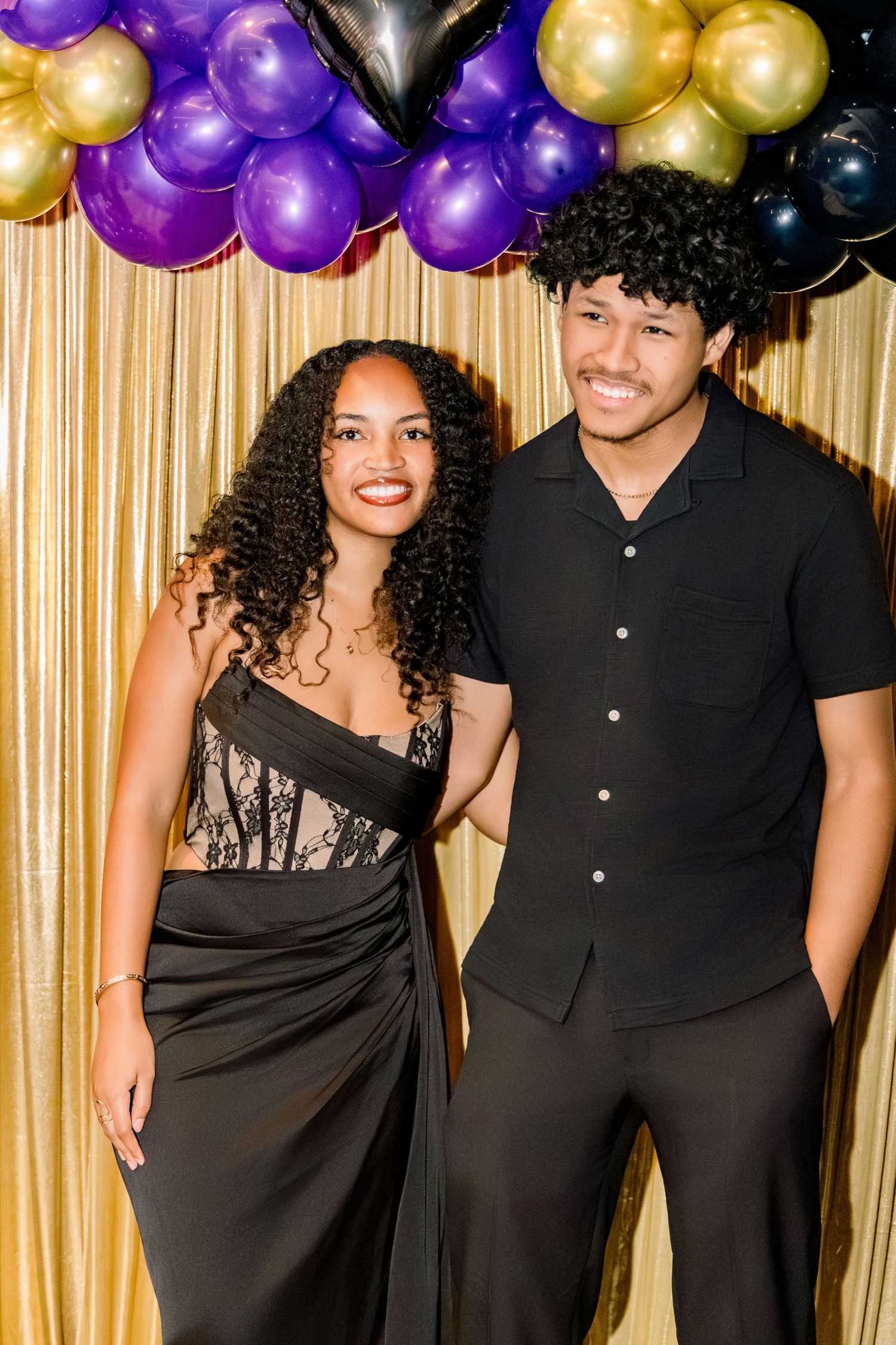 A young couple dressed in elegant black outfits posing together at a prom event with gold and purple balloon decorations.
