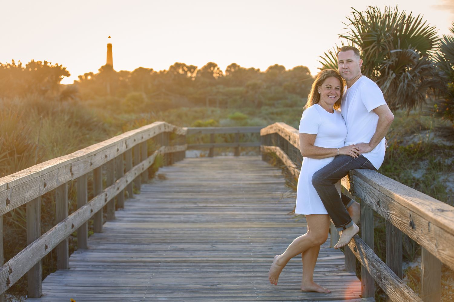 Couple posing on a wooden boardwalk at golden hour with a lighthouse and palm trees in the background.