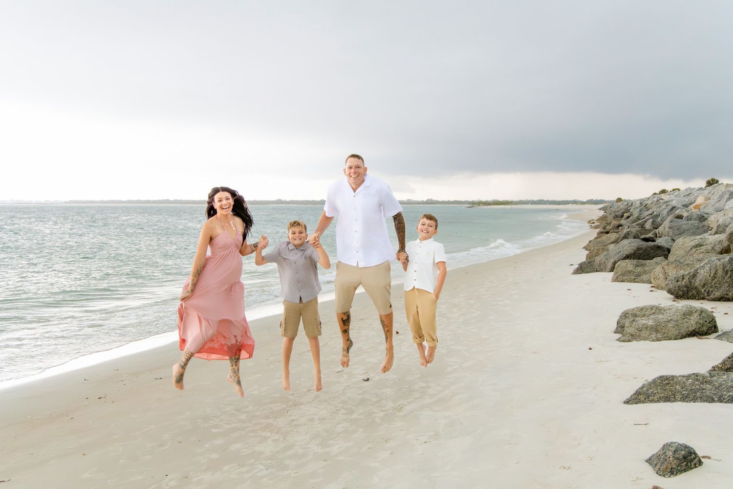 Family of four joyfully jumping on a sandy beach at sunset, with rocky shoreline and ocean waves in the background.