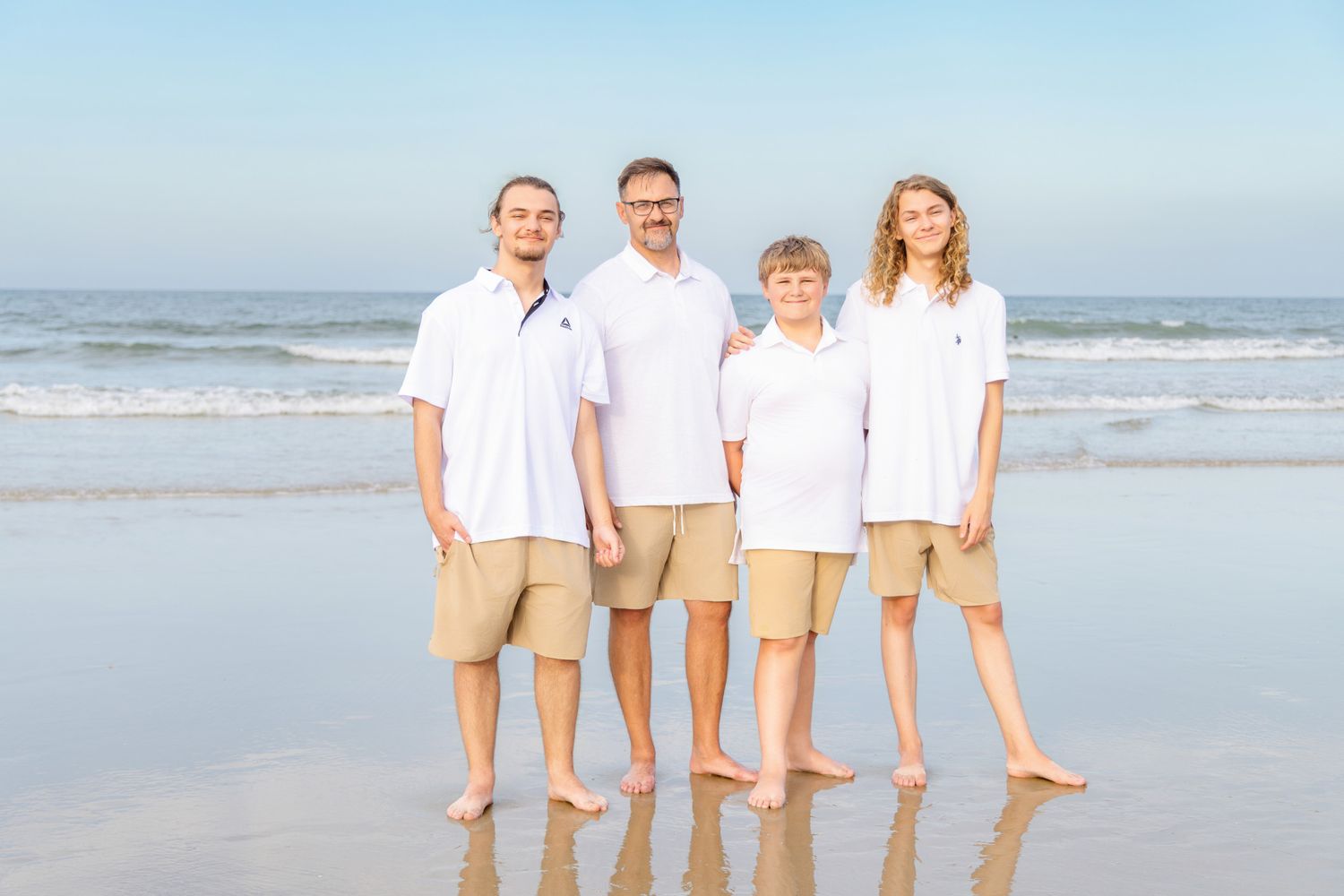 Family of four in matching white shirts and khaki shorts posing on a sunny beach with ocean waves behind them.