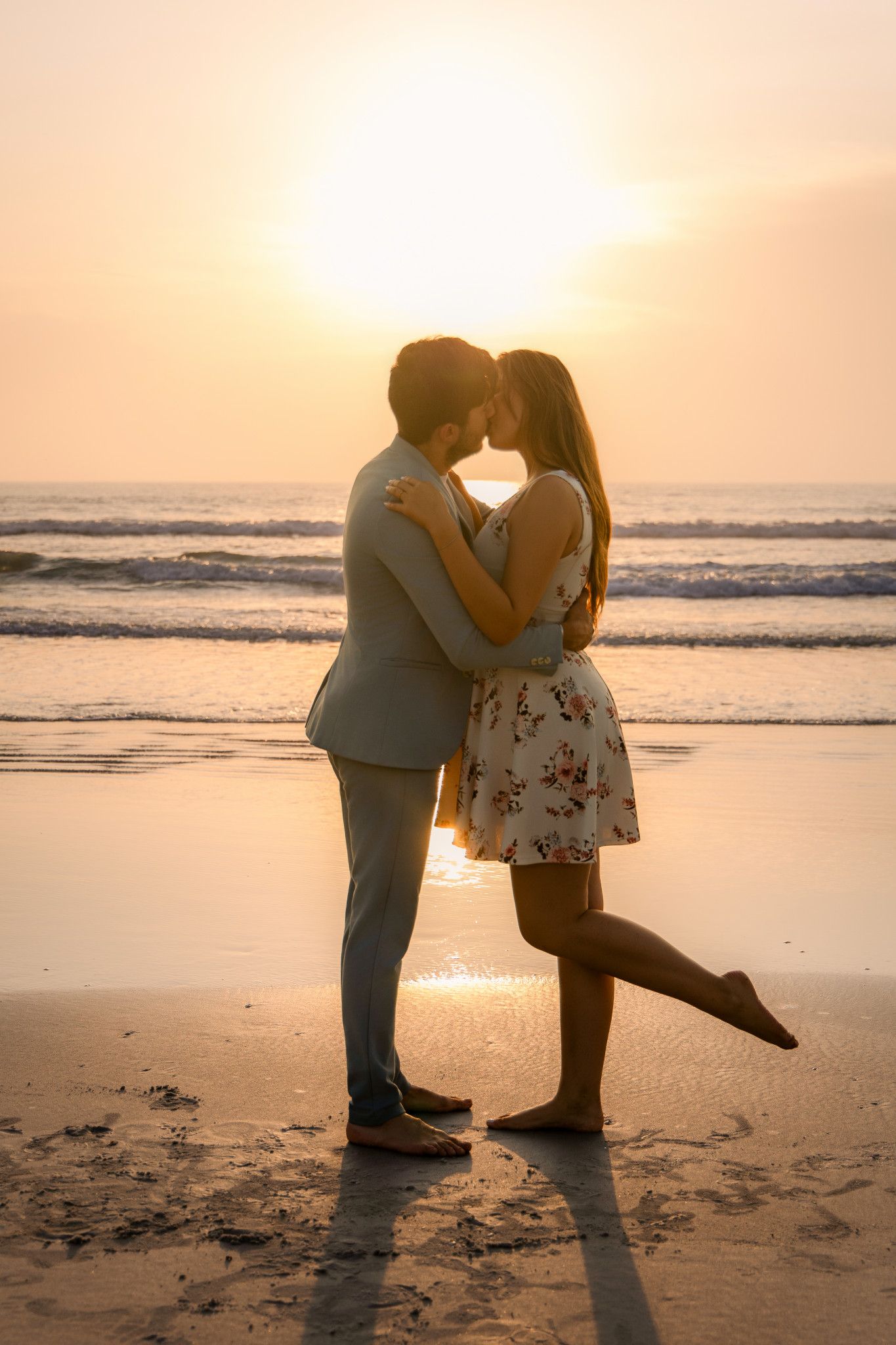 Romantic couple kissing on a beach at golden sunset, woman in floral dress with leg raised, ocean waves in background.