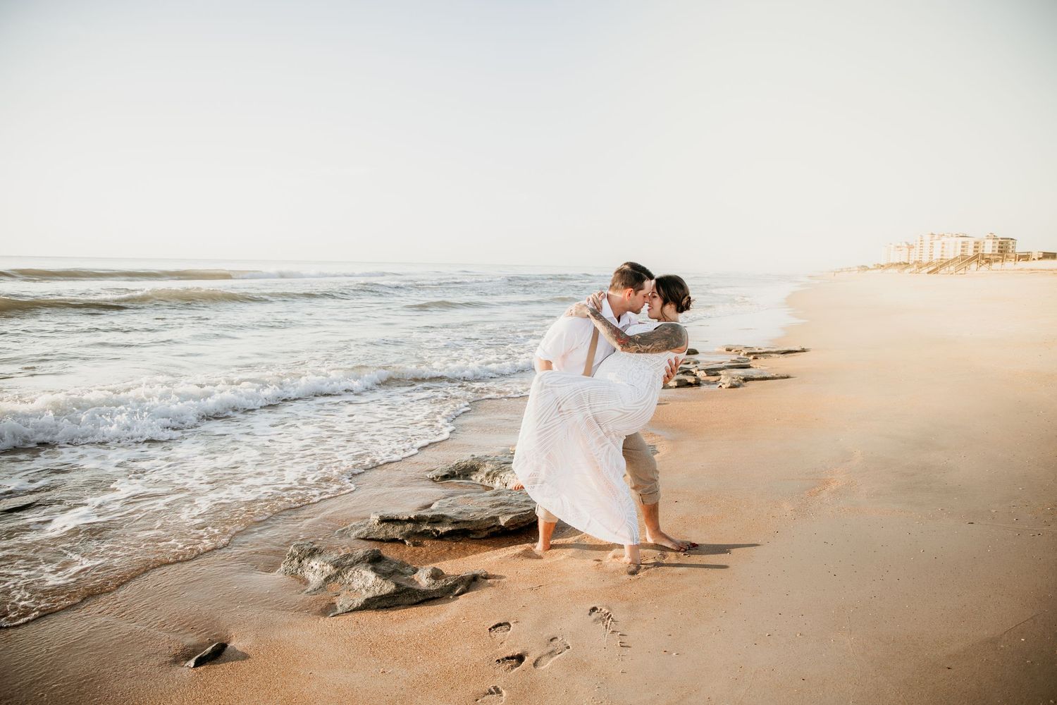 Couple sharing romantic kiss on sandy beach shoreline during golden hour at their beach wedding.