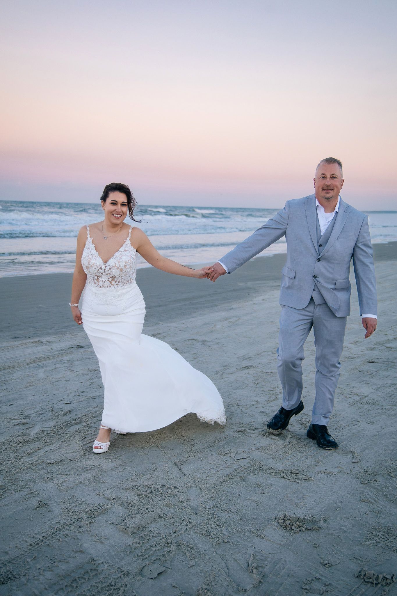 A couple in wedding attire walking along a beach at sunset with the ocean waves in the background.