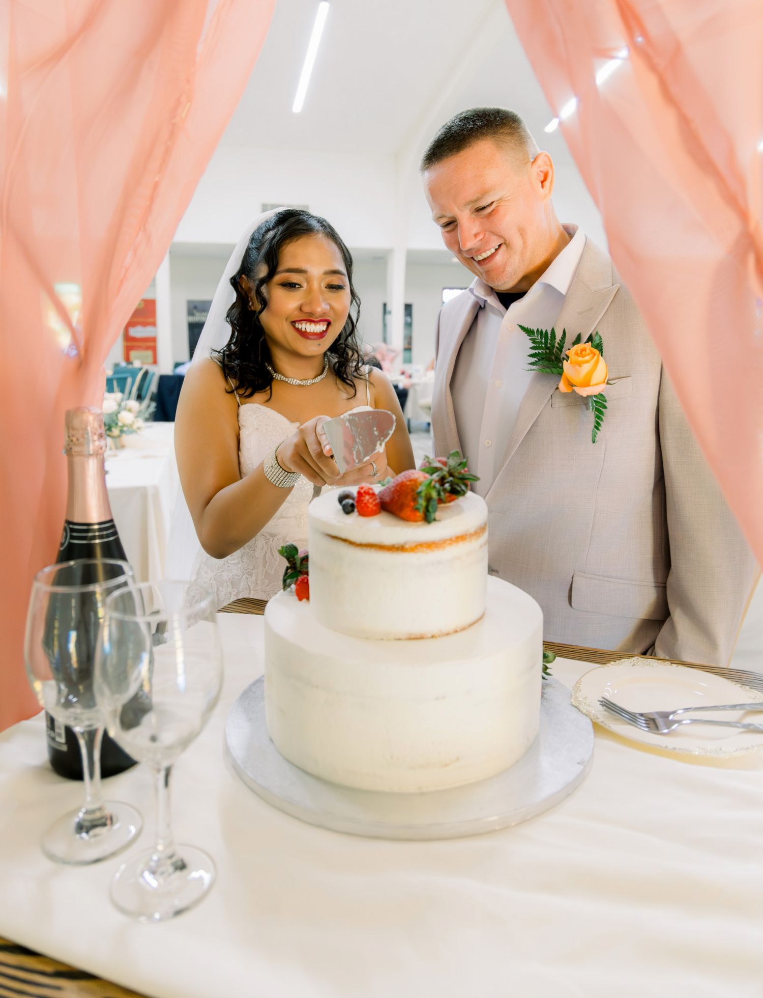 Bride and groom smiling while cutting a two-tier white wedding cake decorated with fresh berries at their reception.