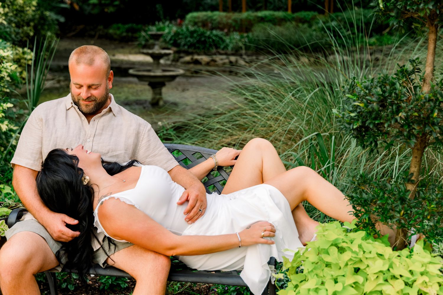 Pregnant woman in white dress reclines on garden bench with partner in lush green outdoor setting.