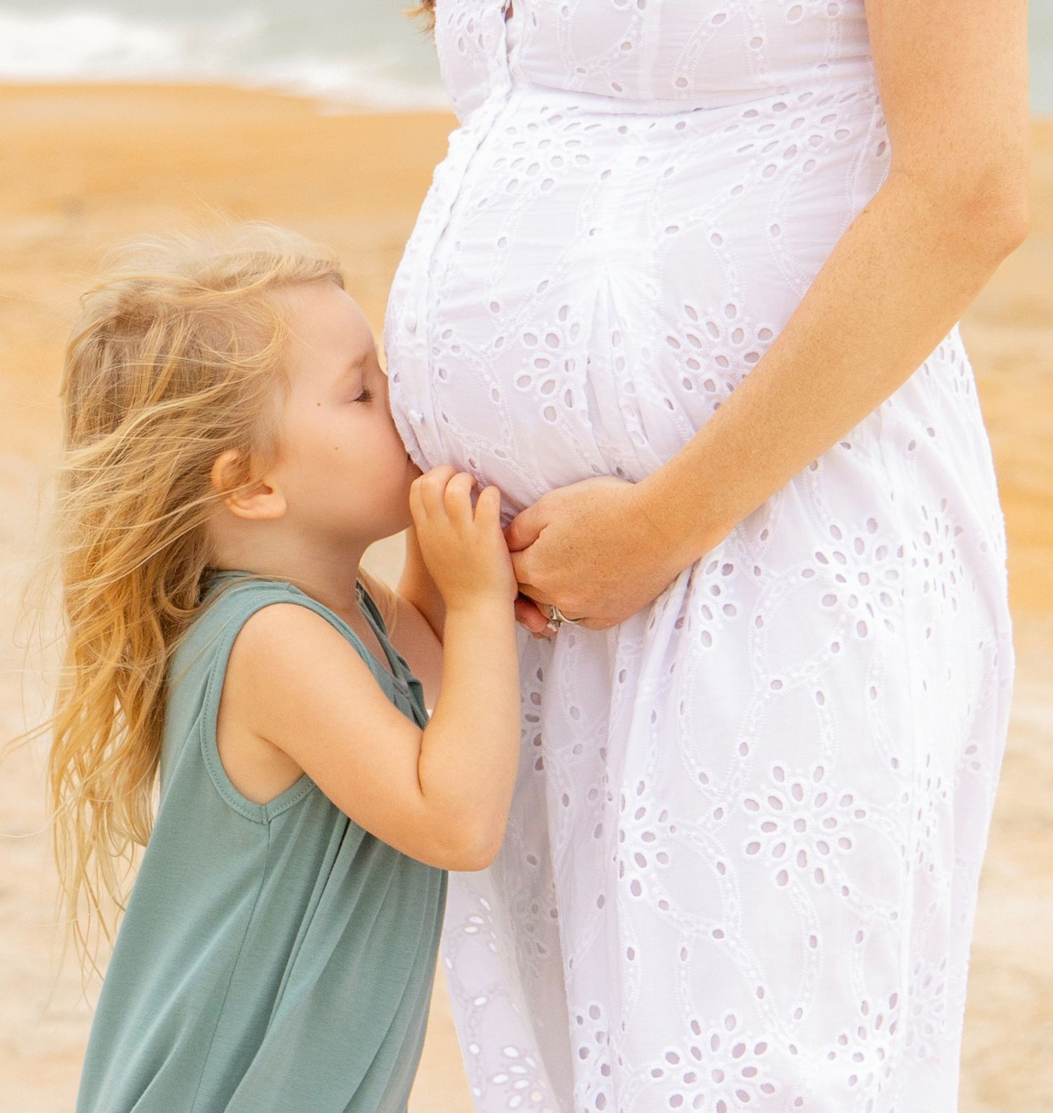 Young girl kissing pregnant mother's belly on a sunny beach, wearing a teal dress beside white eyelet maternity gown.