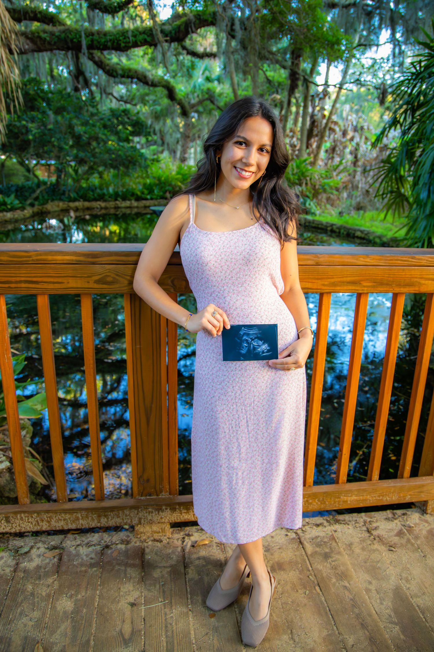 Pregnant woman in pink floral dress holding ultrasound photo on wooden bridge surrounded by lush greenery.