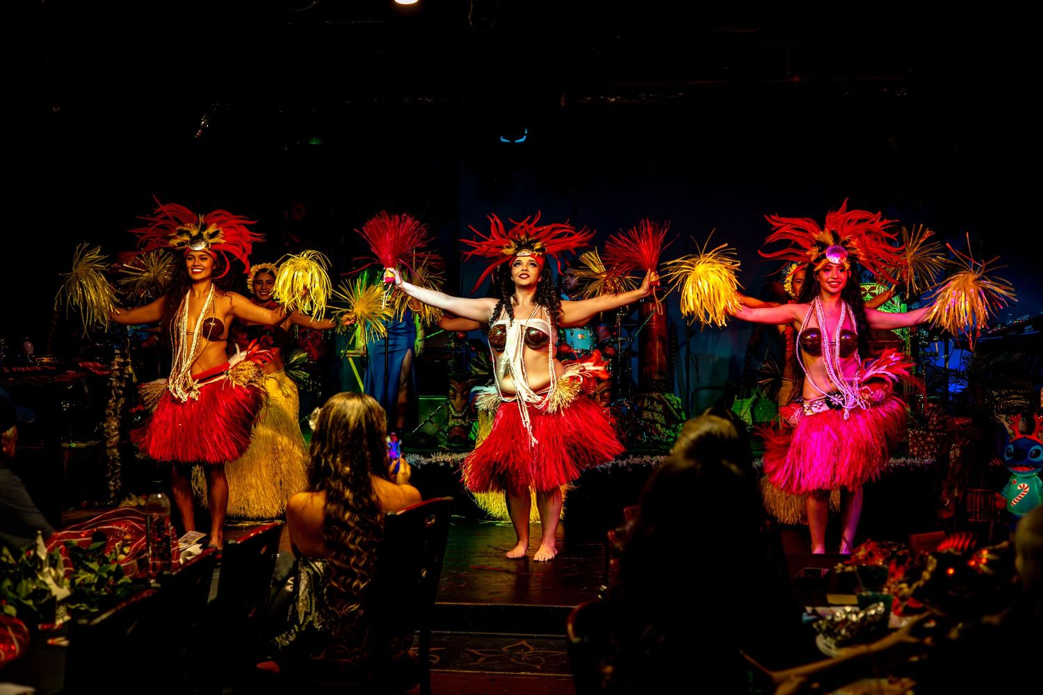 Performers in colorful costumes dance and entertain on a dimly lit stage during a theatrical production.