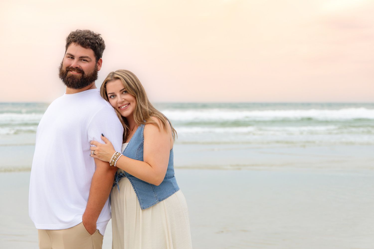 Couple embracing on a beach at sunset, woman in blue top and white skirt smiling at camera.