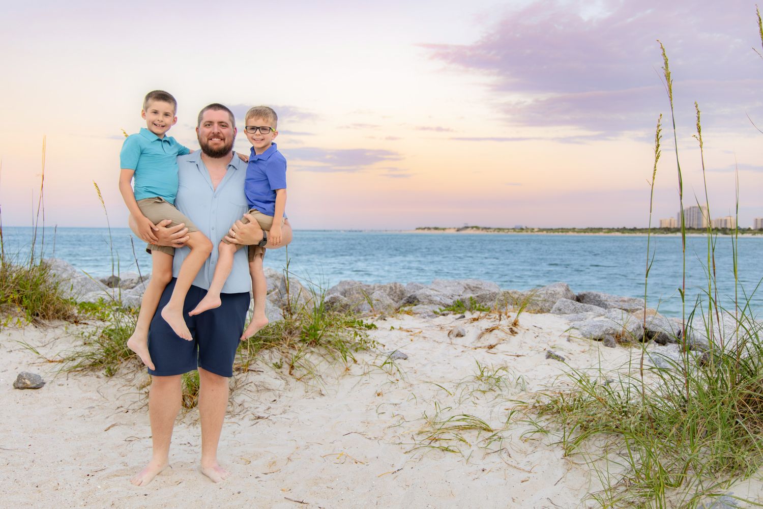 A father holds two young boys on a sandy beach at sunset with turquoise water and sea oats in the background.