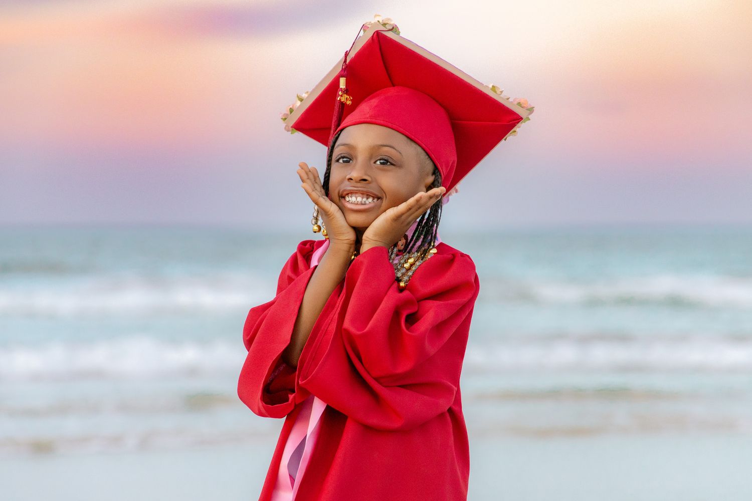 Young graduate in red cap and gown smiling joyfully on a beach at sunset with ocean waves in the background.