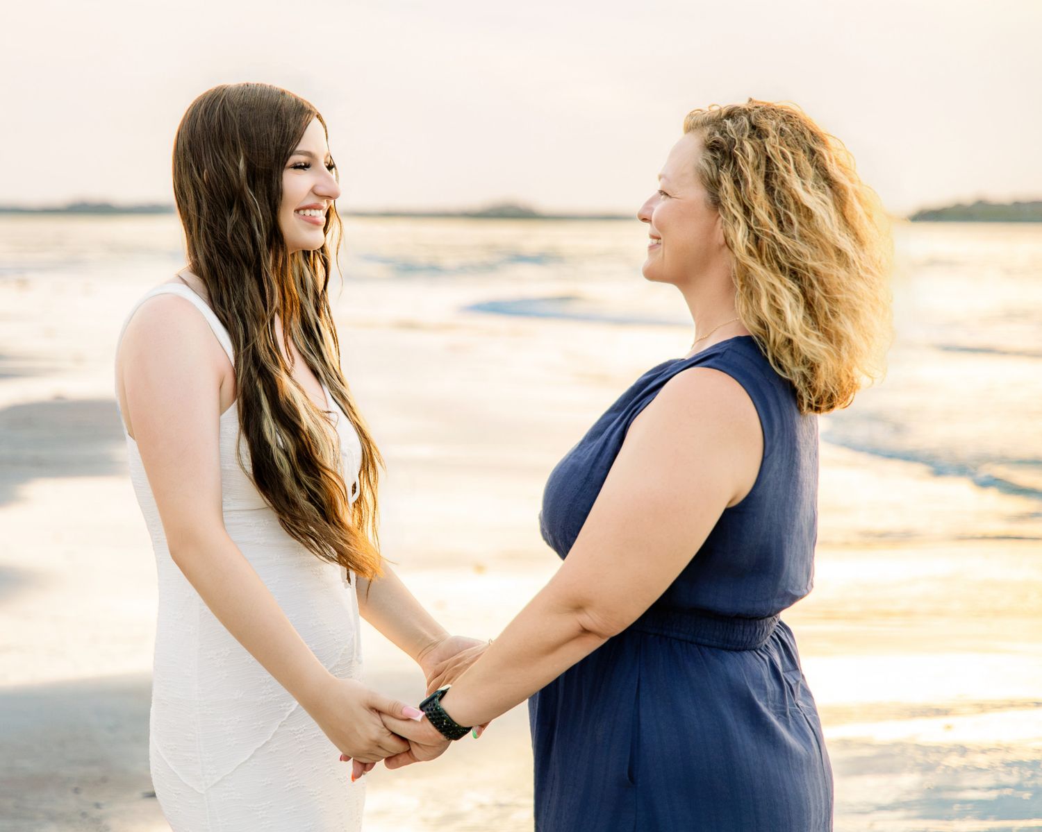 Two women holding hands on a beach at sunset, one in white dress, one in navy, smiling at each other.