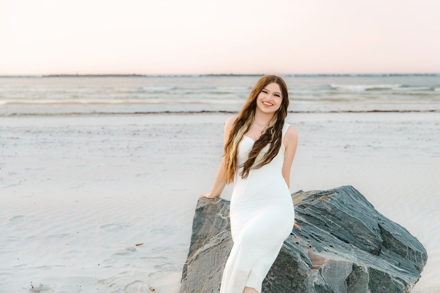 Young woman in white dress posing on beach rocks at sunset with ocean in background.