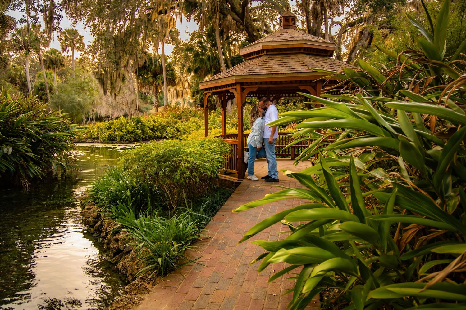 Scenic garden path leads to a wooden gazebo surrounded by lush tropical plants and a peaceful pond at sunset.