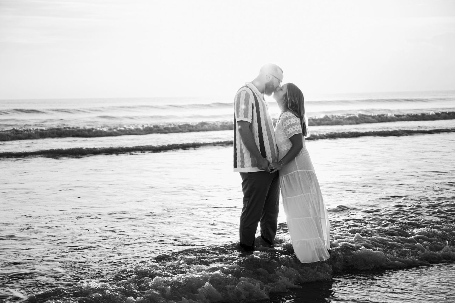 A romantic black and white beach scene with a couple embracing at the water's edge during sunset.