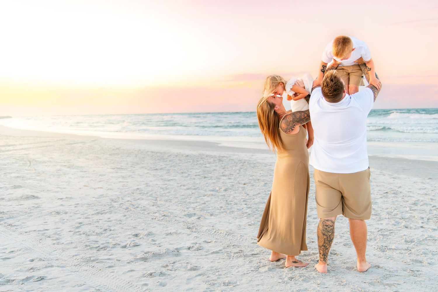 Family of four enjoying a beautiful sunset on the beach, parents holding young children while facing the ocean.