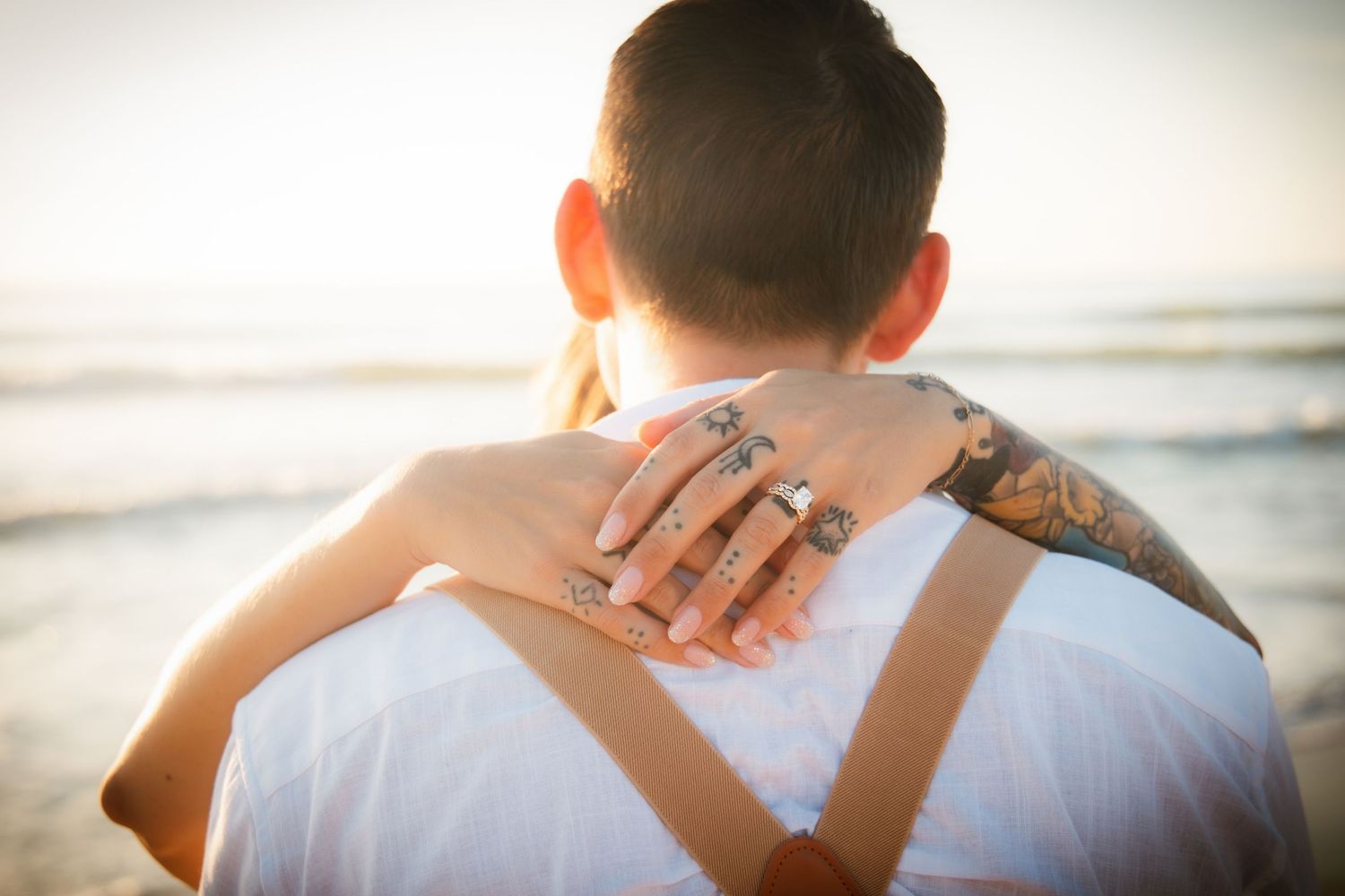 Couple embracing at beach sunset, woman with tattoos and rings on hand resting on man's shoulder.