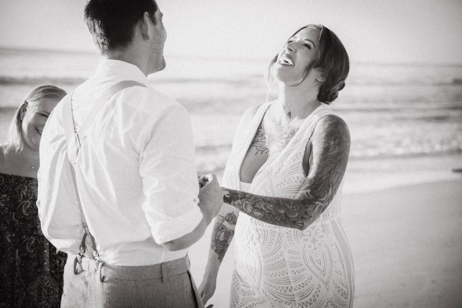 Black and white photo of laughing tattooed bride holding groom's hands on the beach.