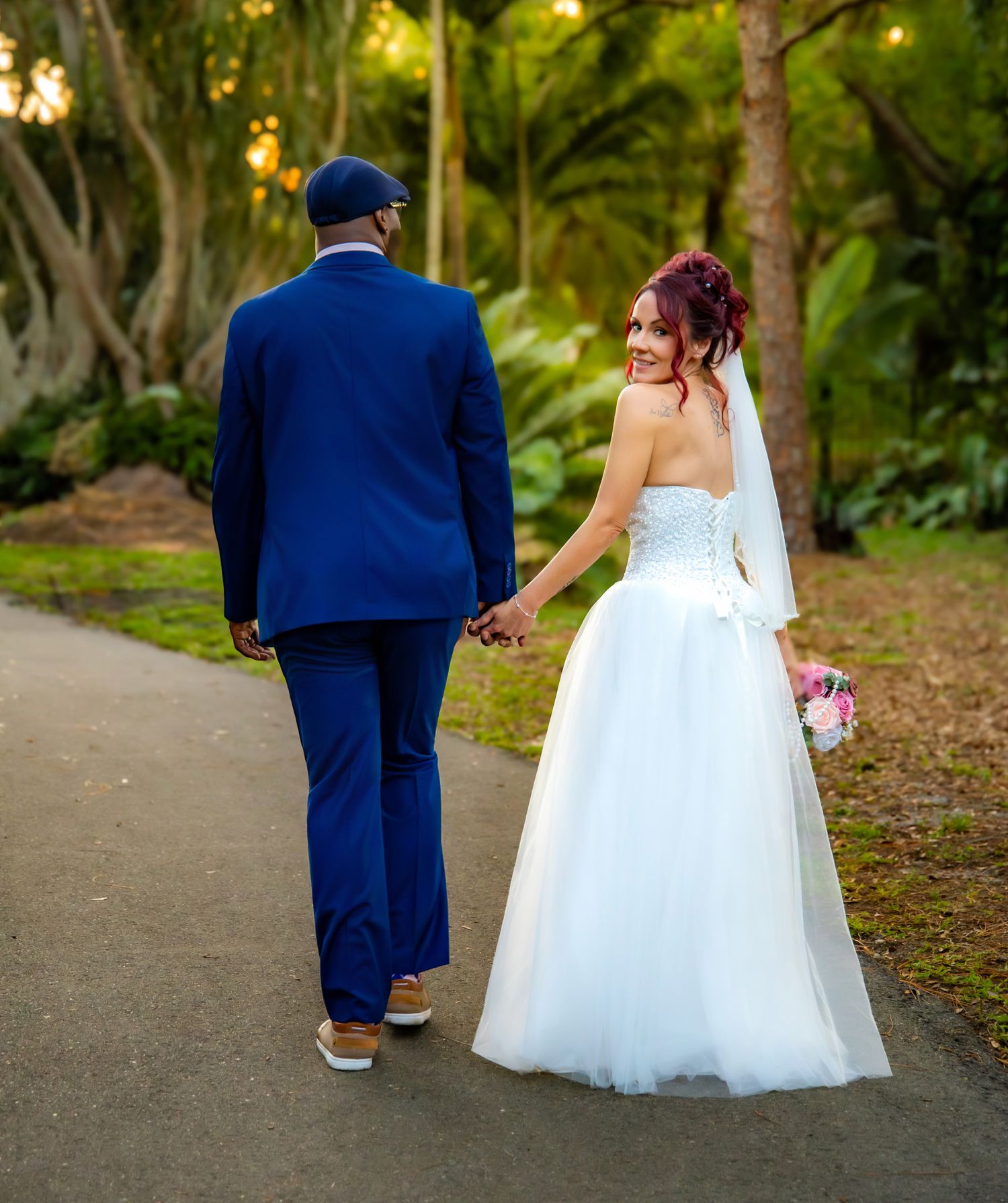 A couple in wedding attire walking down a garden path, with the bride in white and groom in navy blue.