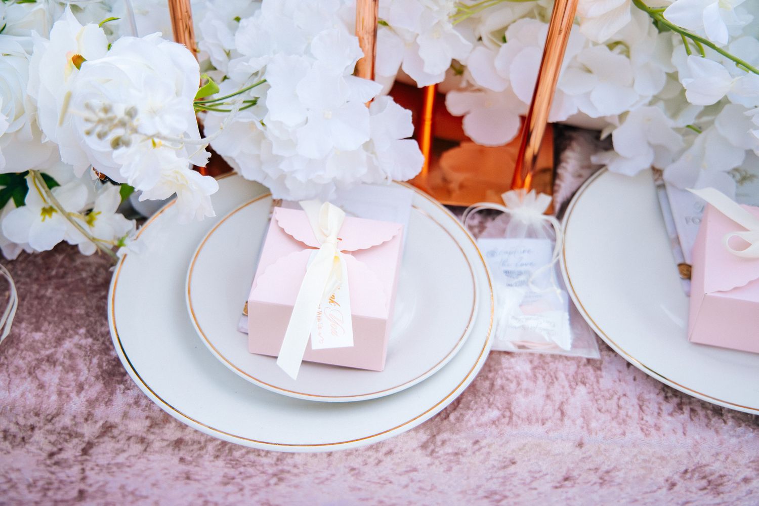 Elegant white floral centerpieces with pink gift boxes on white plates create a romantic table setting.