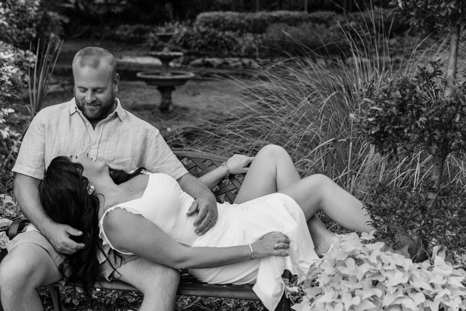 Black and white maternity photo of couple relaxing outdoors, pregnant woman lying down with partner beside her in garden.