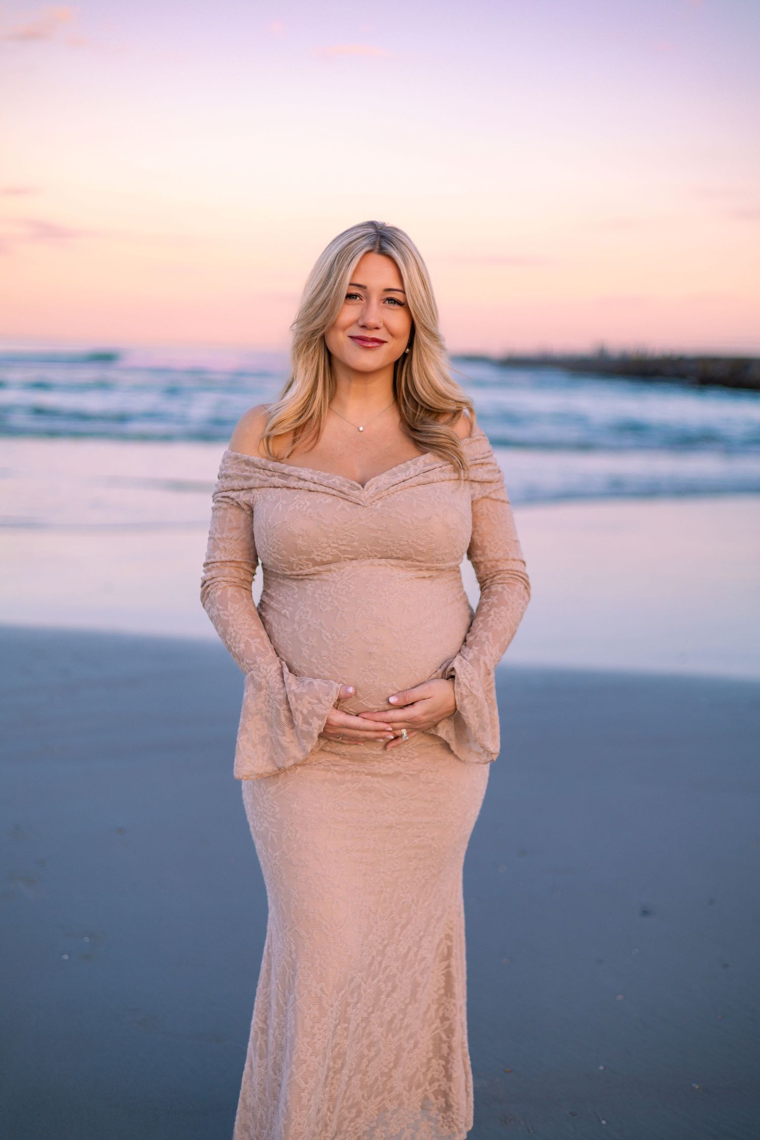 A woman in a beige lace dress poses on a beach at sunset with soft pink and purple sky in the background.