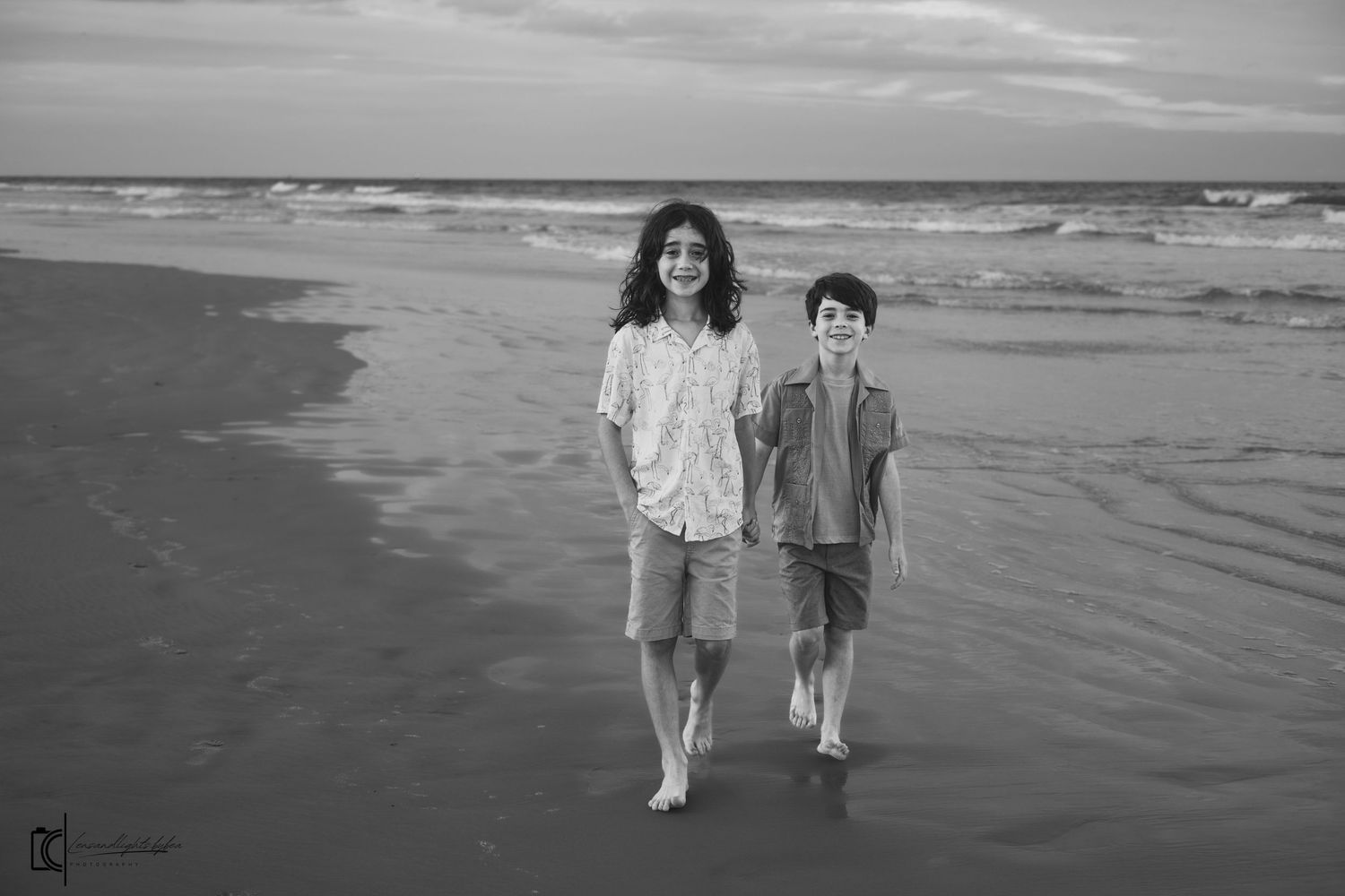 Black and white photo of children walking along a beach shoreline with waves and sand visible.