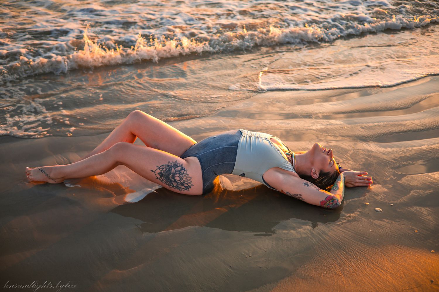 A figure lies on wet sand at the beach during golden hour as gentle waves wash over them creating a dreamy coastal scene.