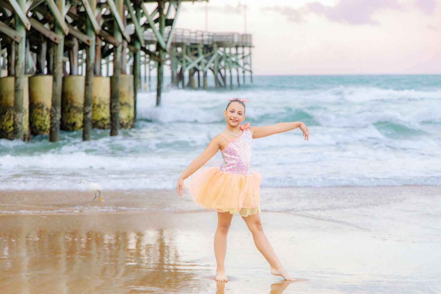 Young girl in pink tutu dancing joyfully on beach near pier with ocean waves in background.