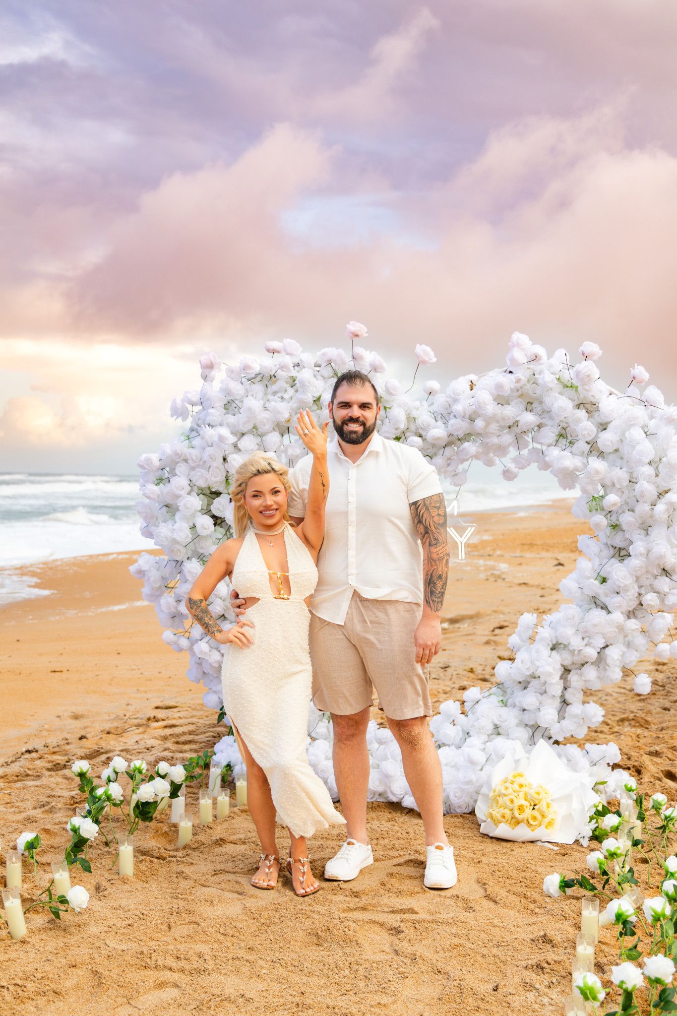 Smiling couple on a beach at sunset posing under a white floral heart arch as the woman shows her engagement ring.