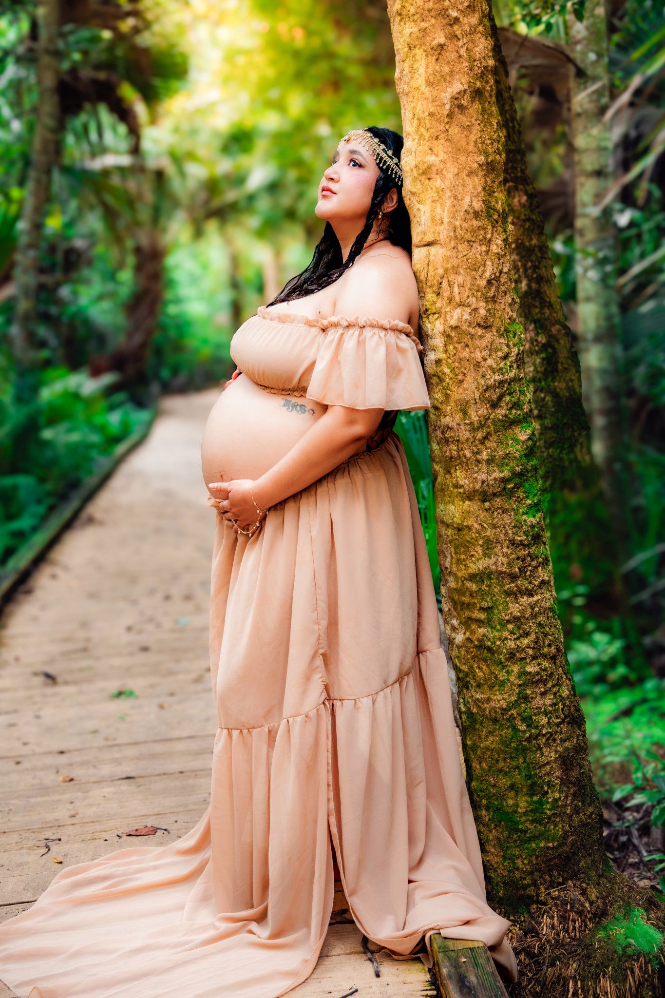 Pregnant woman in a peach off-shoulder maxi dress leaning against a tree on a tropical boardwalk path.