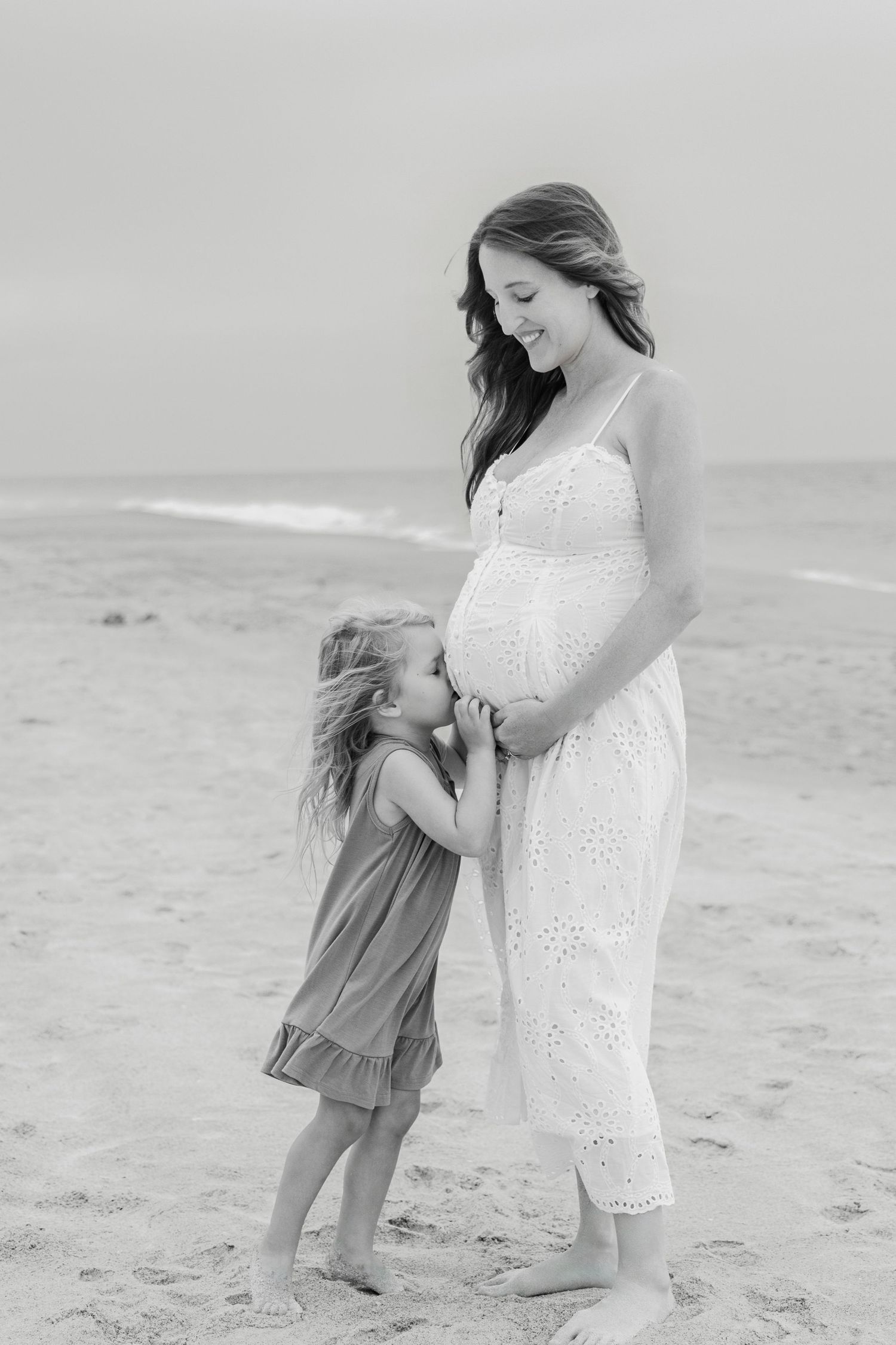 Black and white photo of a pregnant woman on the beach with a young girl kissing her baby bump.
