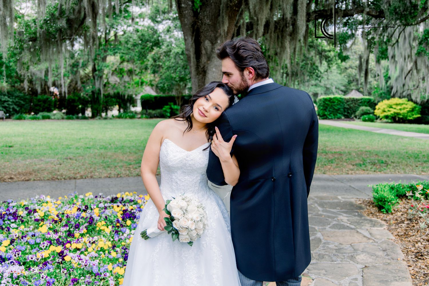 Bride and groom share a tender moment outdoors, surrounded by colorful flowers and Spanish moss-draped trees.