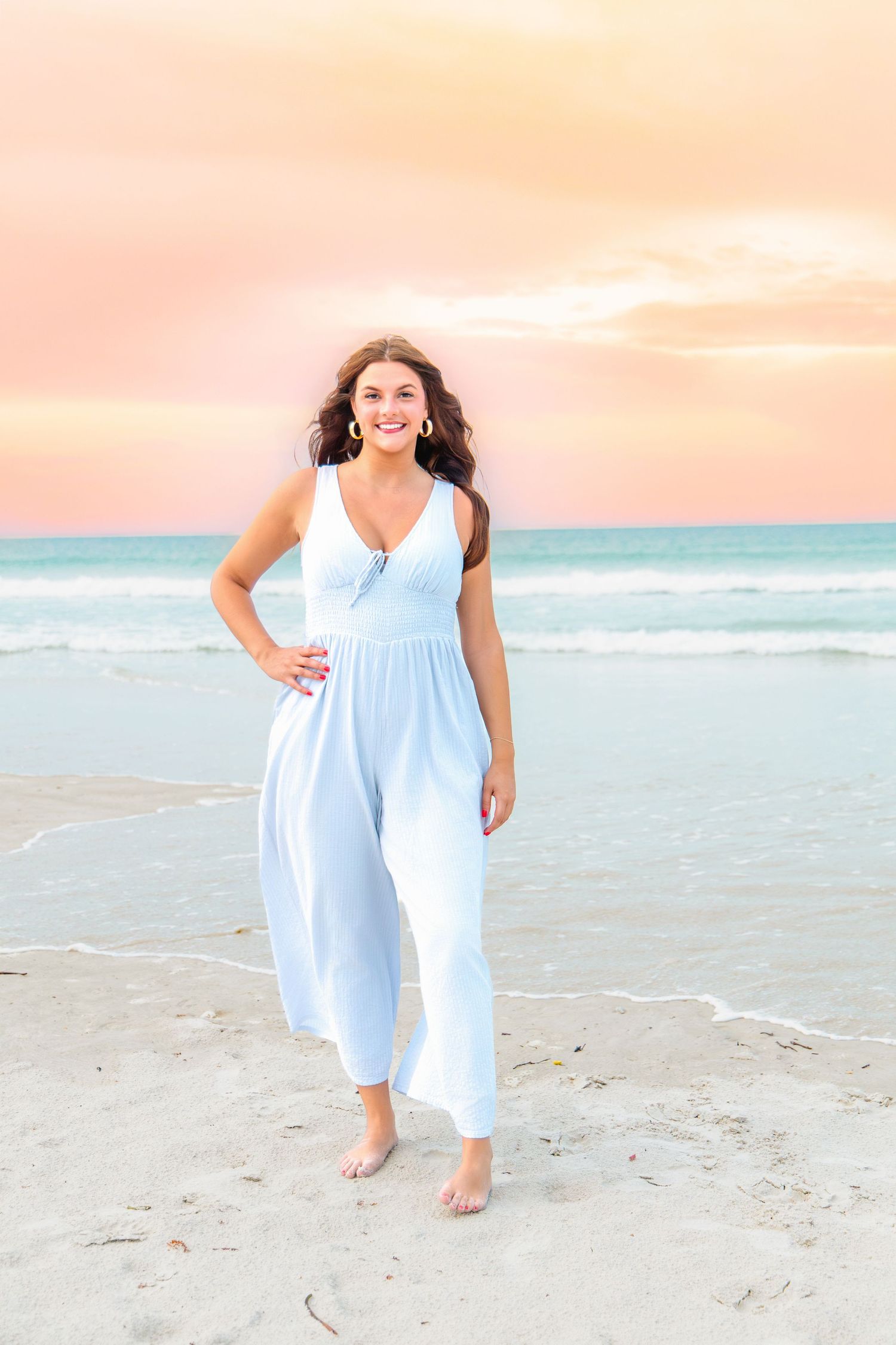 Young woman in light blue jumpsuit poses barefoot on sandy beach at sunset with turquoise ocean waves behind her.