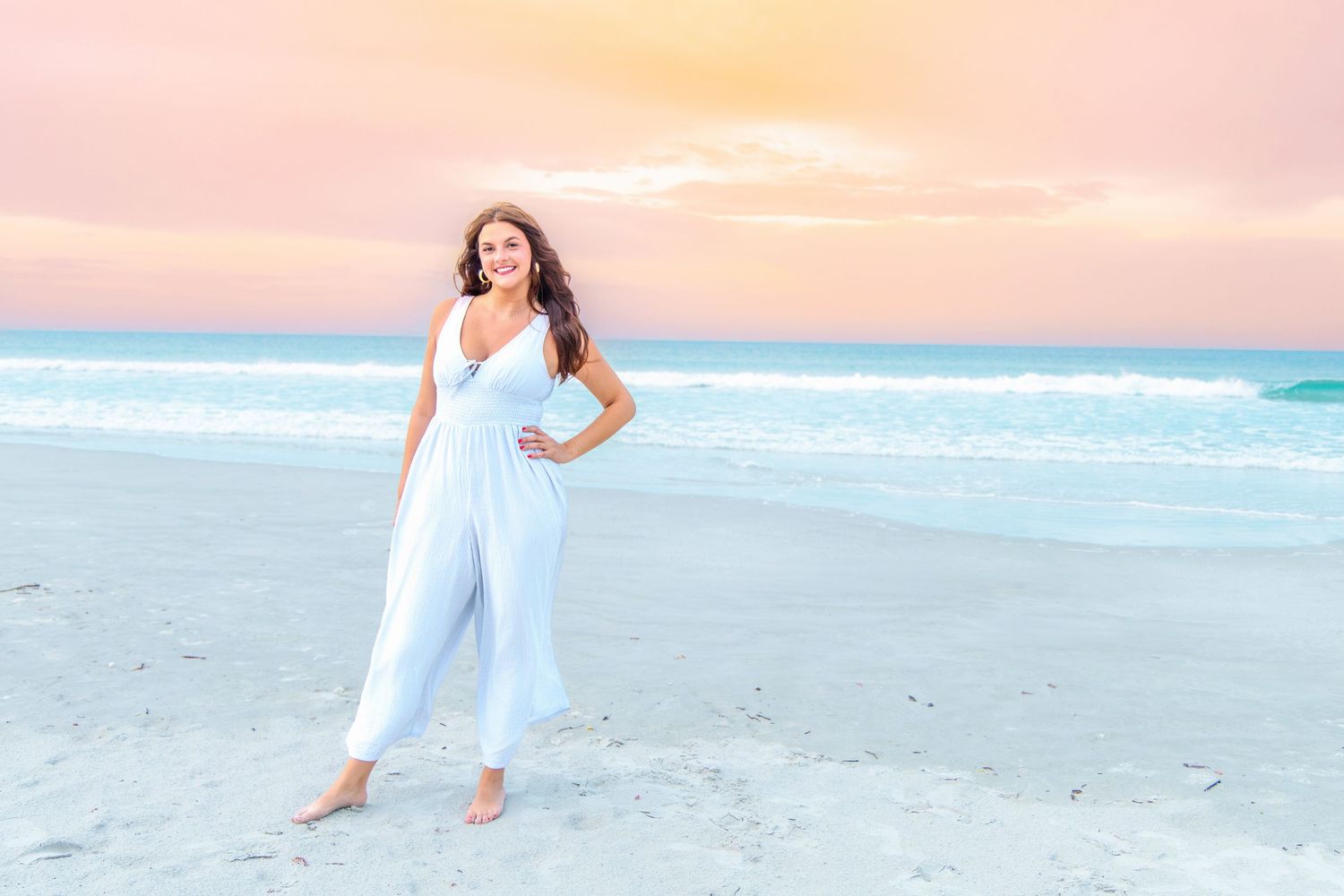 Woman in white jumpsuit posing on a sandy beach at sunset with turquoise ocean waves in the background.