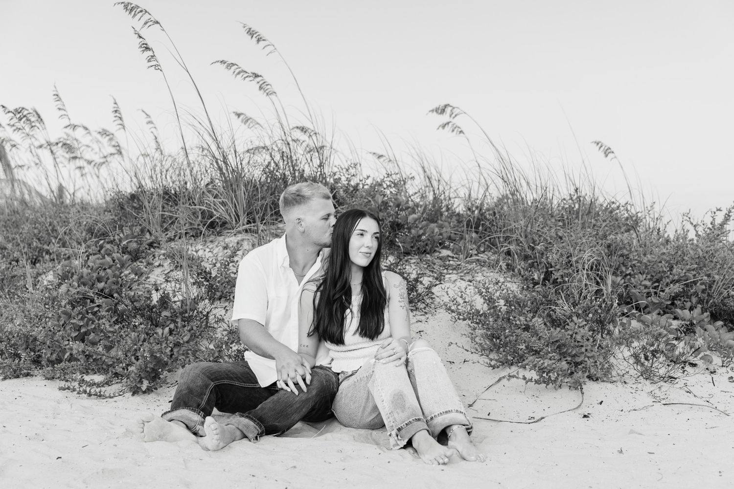 Black and white photo of couple sitting together on beach sand dunes with sea oats in background.