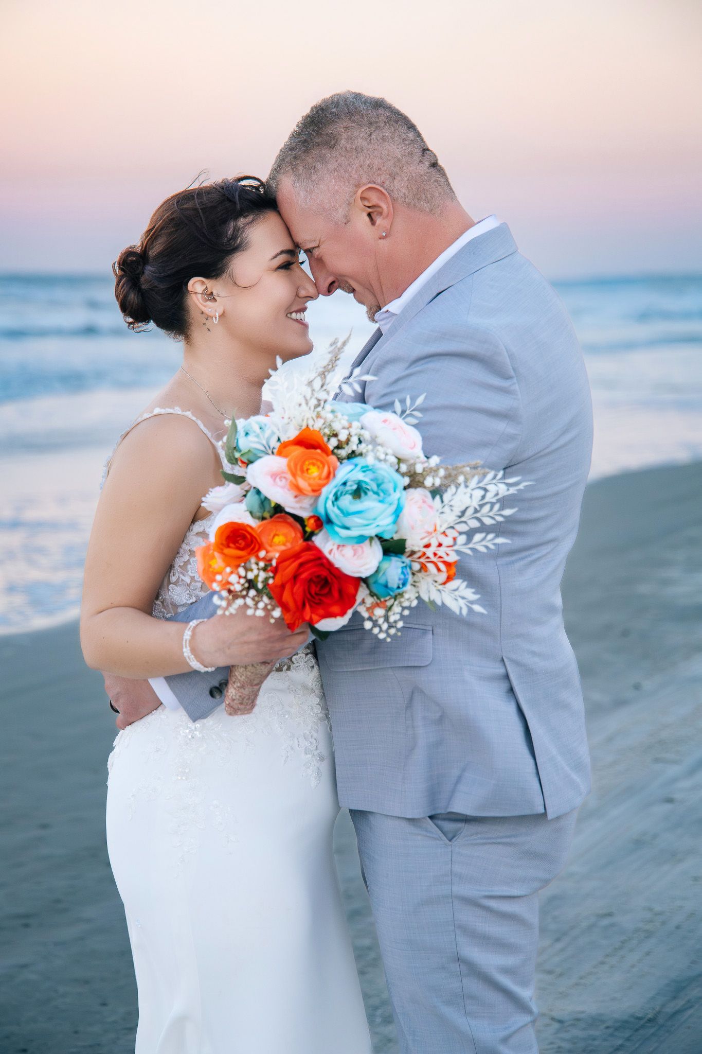 A romantic beach wedding photoshoot at sunset featuring a couple in grey suit and white dress with orange and blue bouquet.