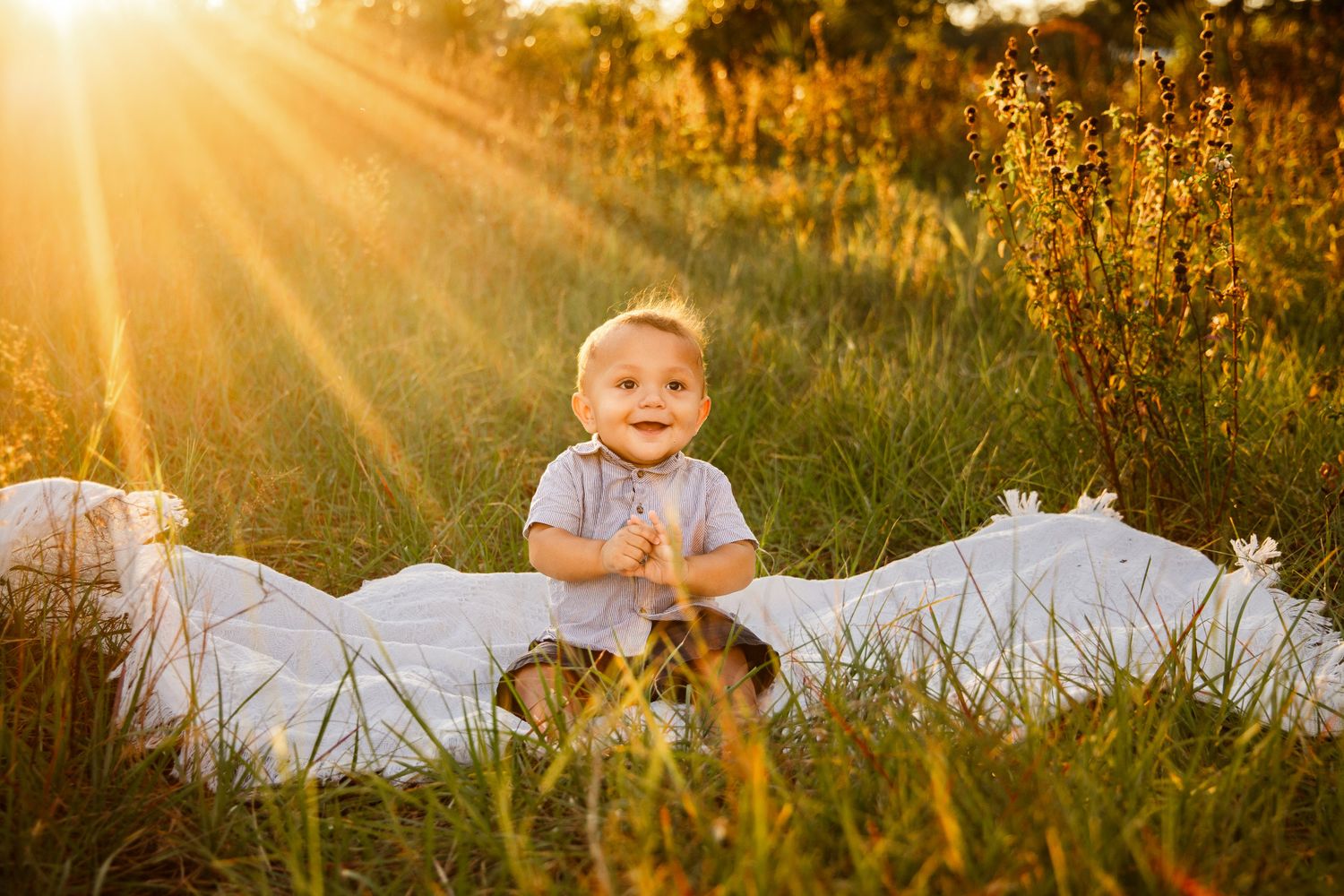 Smiling baby in a gray shirt sits on a white blanket in a grassy field as golden sunset light streams through the trees.