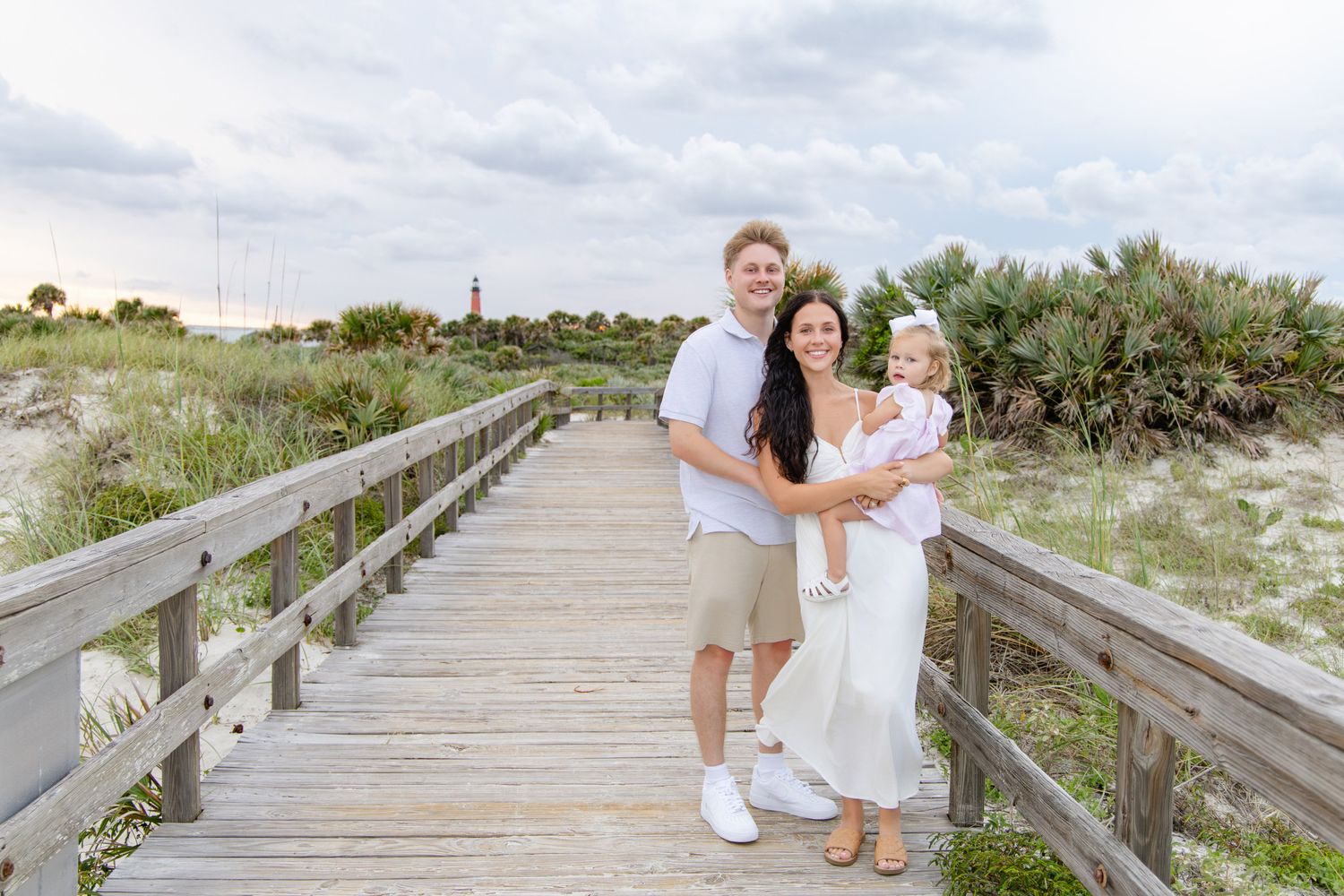 Family of three smiling on a wooden beach boardwalk, parents holding toddler, dressed in white and khaki tones.
