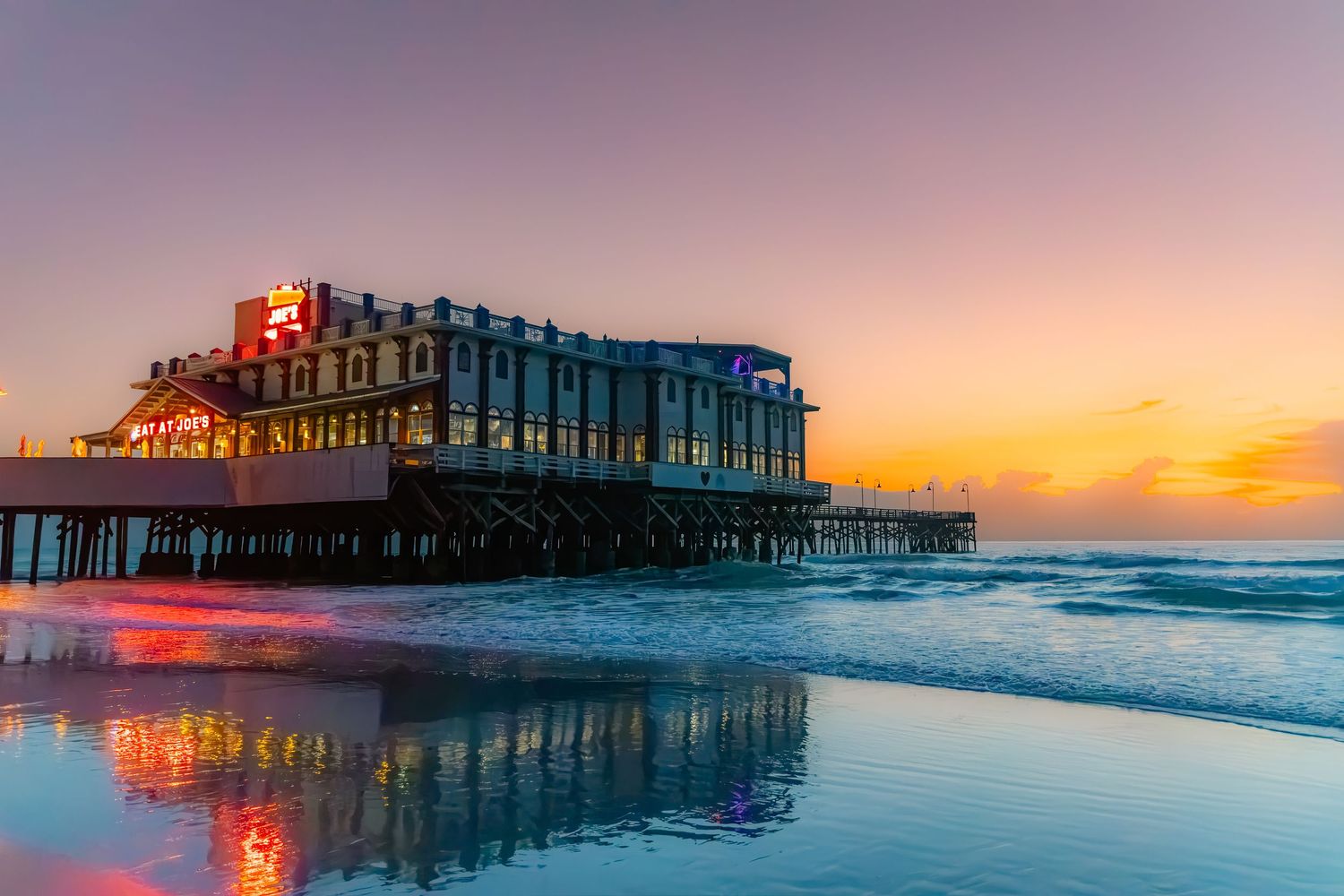 A stunning pier at sunset with glowing lights reflecting on wet sand, surrounded by colorful sky over calm ocean waves.