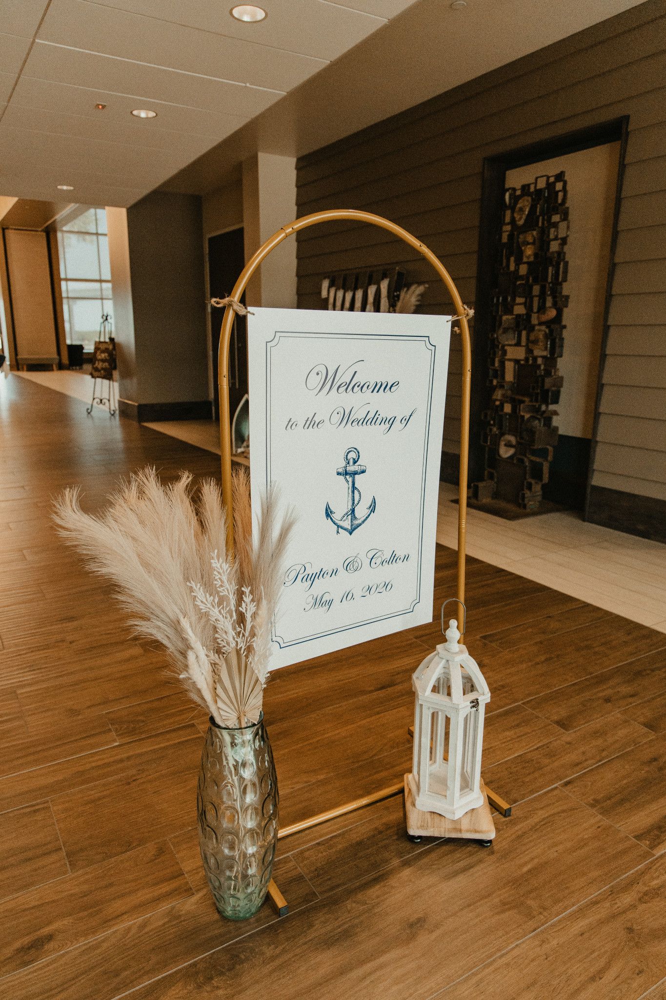 Nautical wedding welcome sign on gold arch with pampas grass and white lantern on hardwood floor.