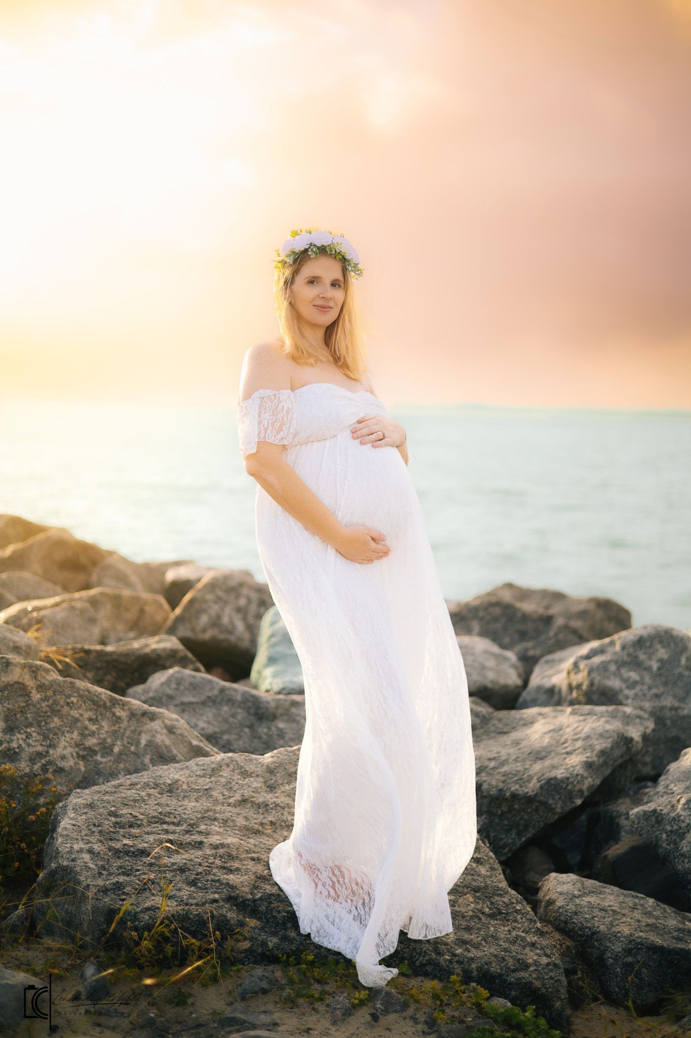 A figure in a flowing white dress poses gracefully on coastal rocks during golden hour sunset.