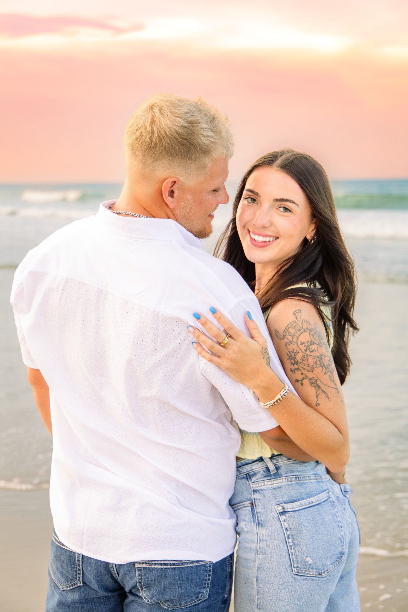 Couple embracing on beach at sunset, woman with dark hair smiling at camera, man in white shirt, ocean backdrop.