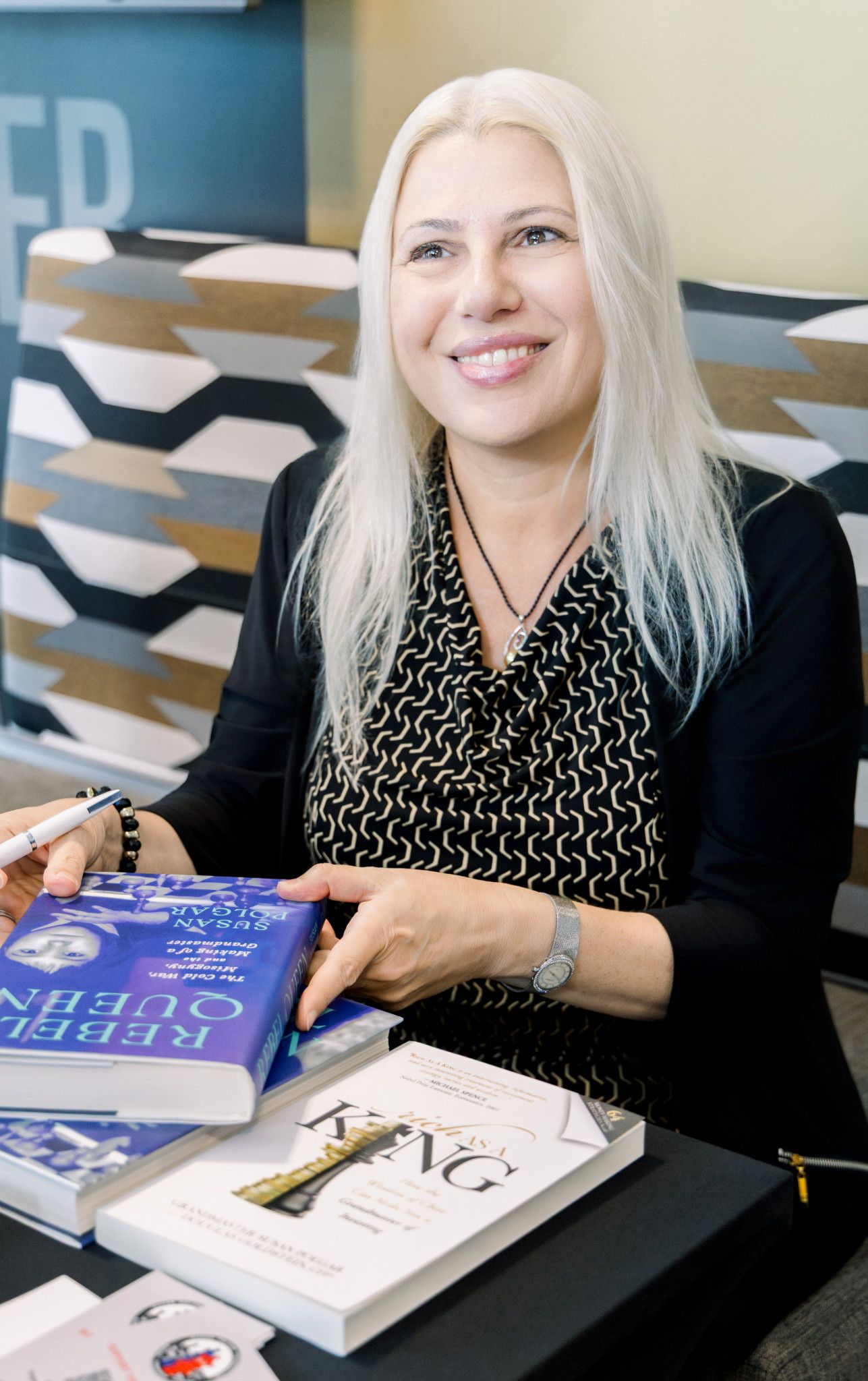 Woman with white hair signing books at a table, smiling at a book signing event.
