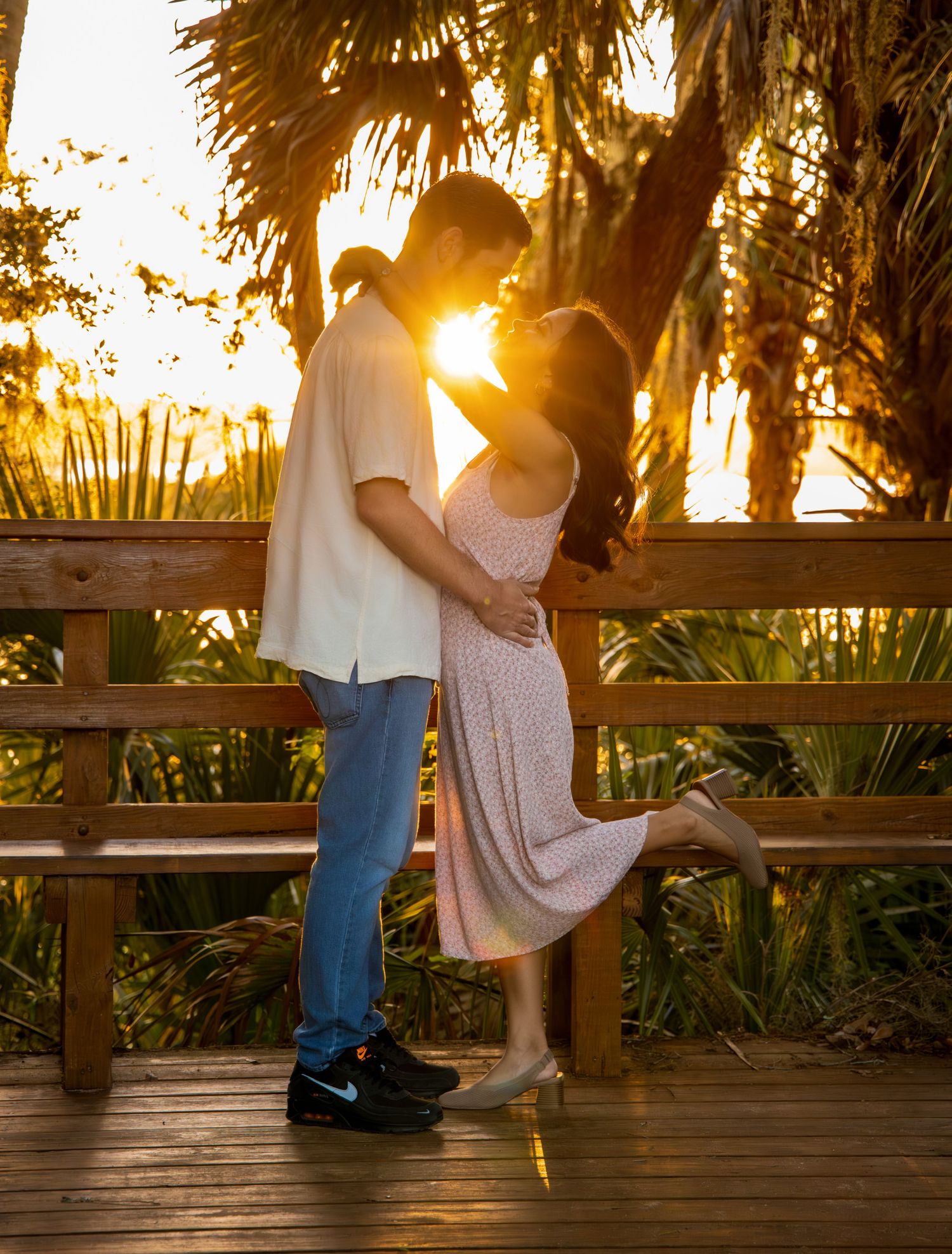 Couple sharing a romantic kiss on a wooden deck at golden sunset, surrounded by lush tropical palm trees.