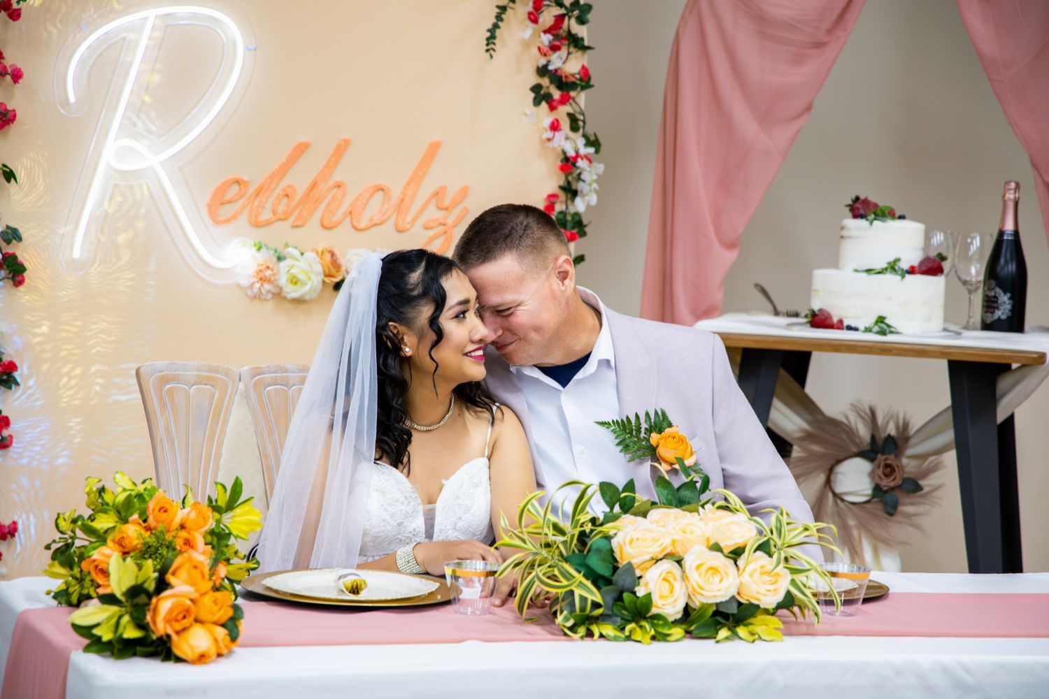 Bride and groom share a romantic moment at floral sweetheart table with neon sign and wedding cake backdrop.